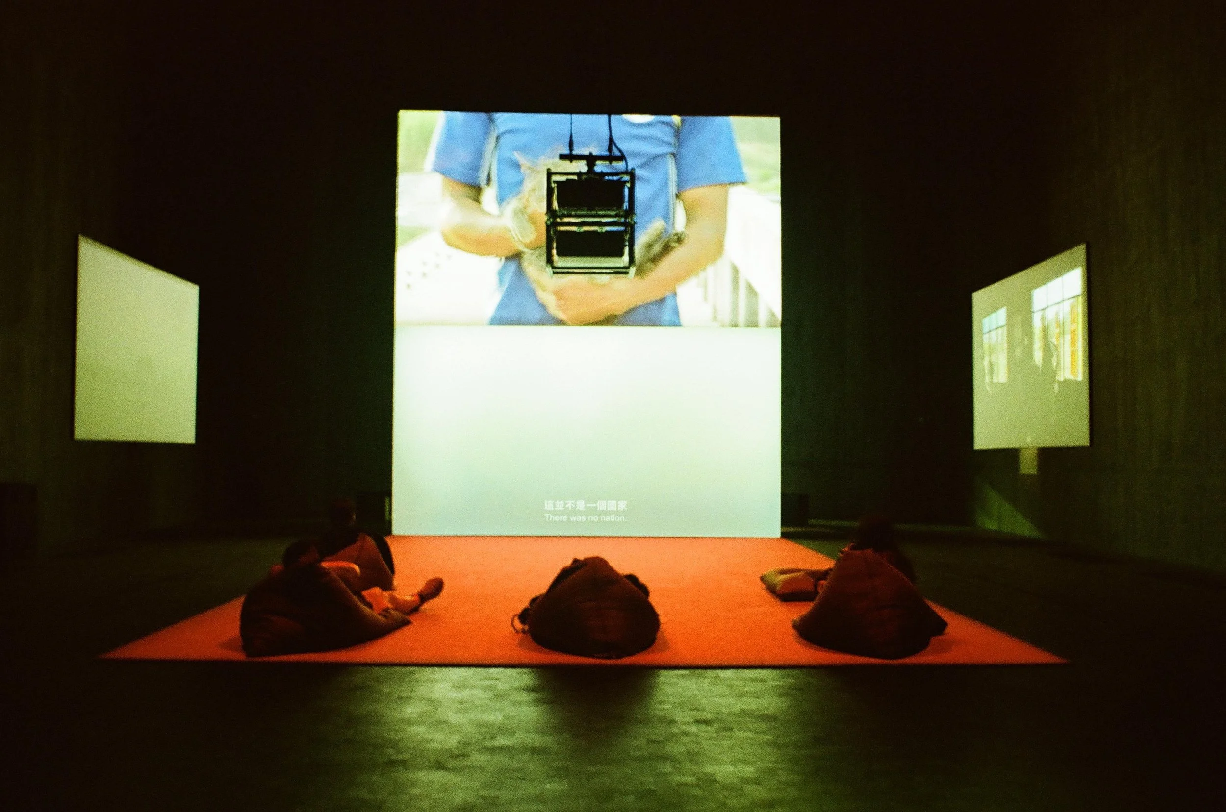 People sitting on a red carpet in front of three screens displaying different images, one with a person holding a camera, and the other two with abstract visuals, in a dark room.