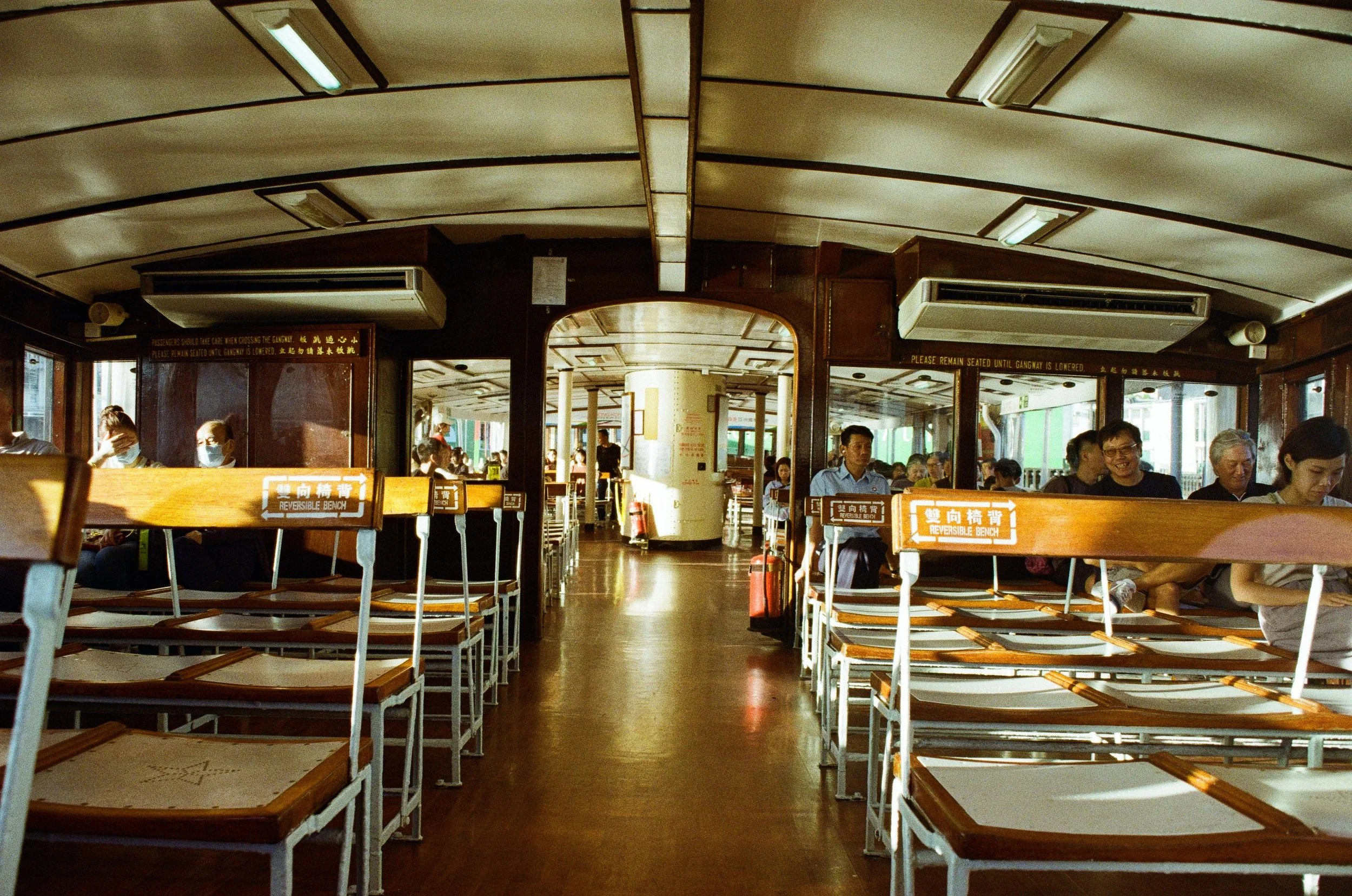 Interior of a ferry boat with empty reversible benches in the foreground and passengers sitting along the sides, some wearing masks, with a view toward the back of the boat and more passengers seated.