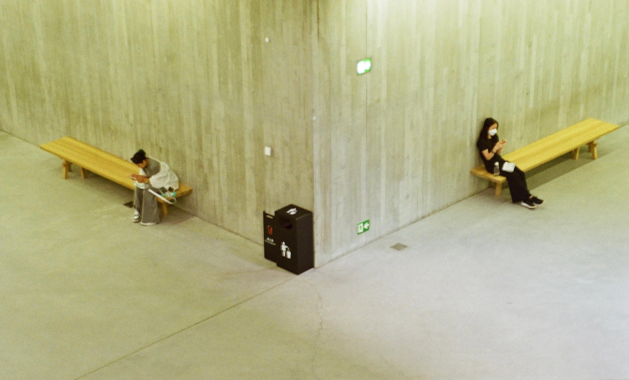 Two women sitting on separate wooden benches in a large, minimalist indoor space, both wearing face masks and engaged with their phones. One woman has a water bottle and notebook beside her.