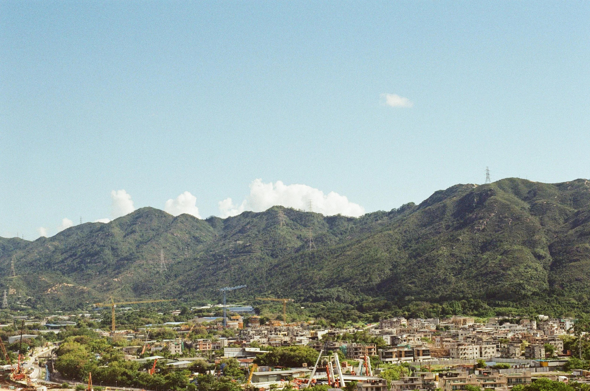 A cityscape with numerous residential buildings in front of green mountains under a clear blue sky with scattered clouds.