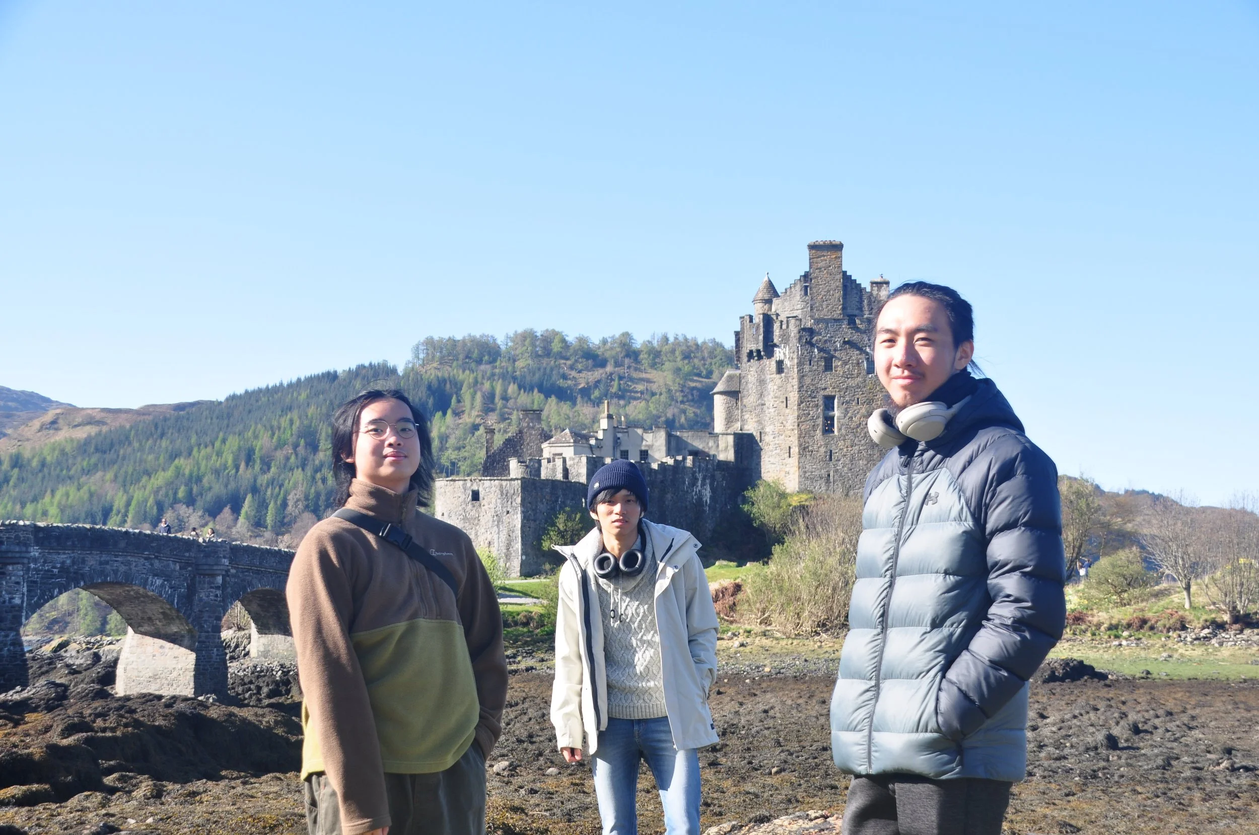 Three young people standing outdoors near a stone castle on a hill with a clear blue sky in the background.