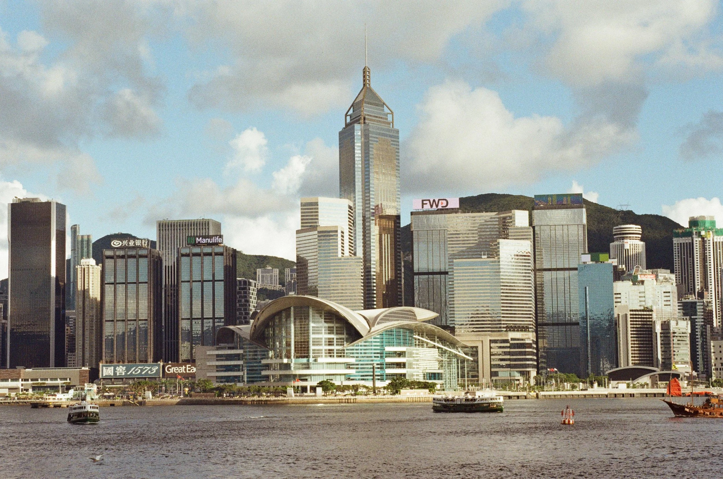 A city skyline with tall modern skyscrapers along a waterfront, including the tallest building with a spire, under a partly cloudy sky.