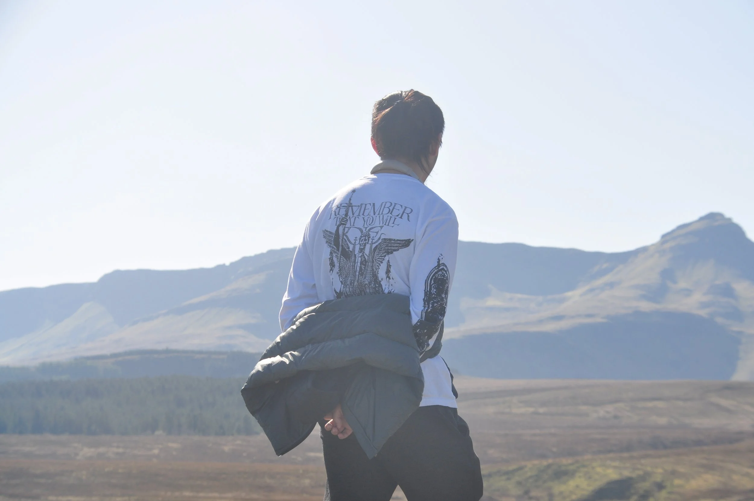 A person standing outdoors in a mountainous landscape with hills and a clear sky, wearing a white shirt and a gray jacket.