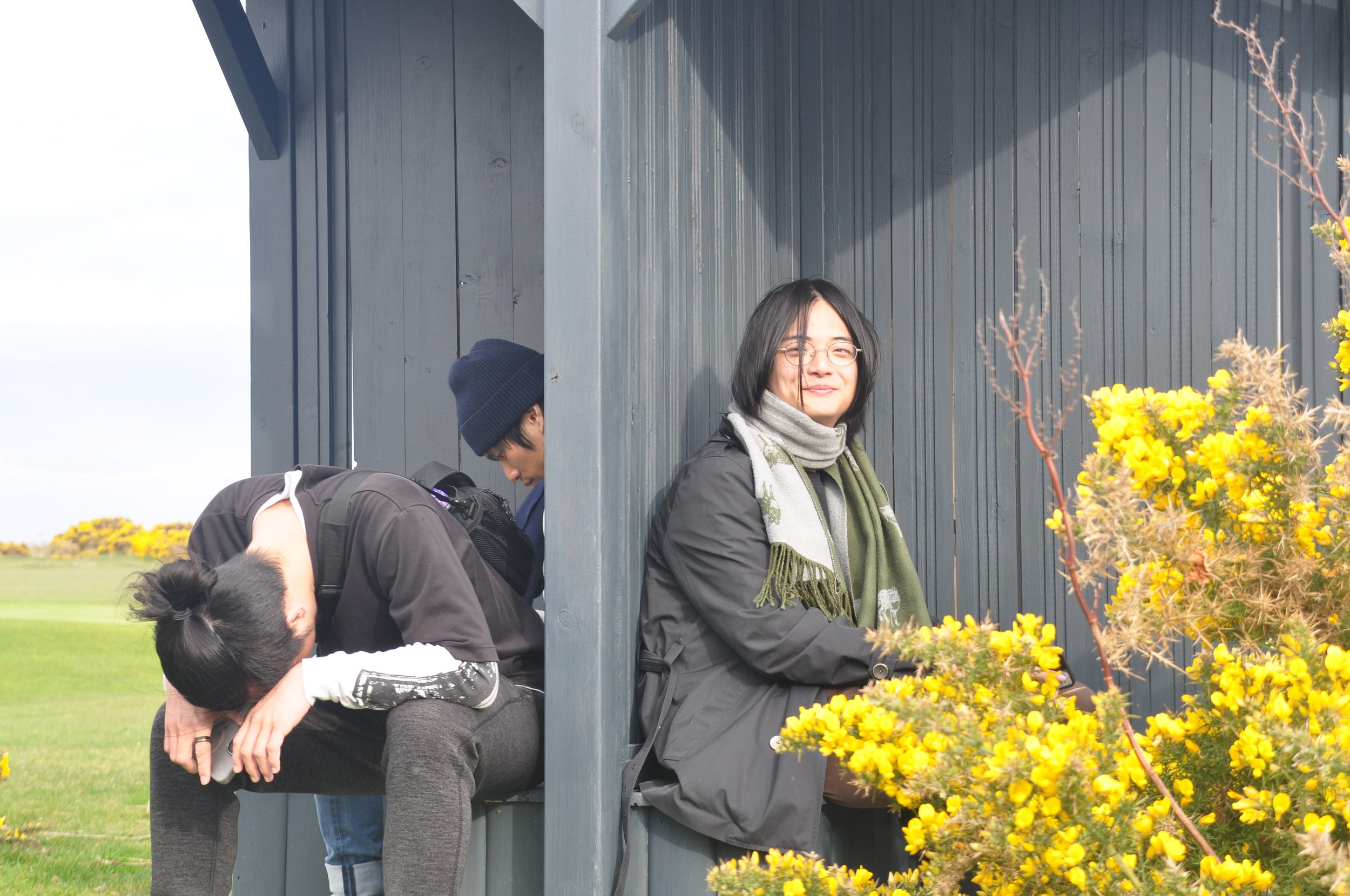 A woman with glasses and shoulder-length hair sitting outside next to a yellow flower bush, smiling at the camera. Behind her, two men are sitting on a bench with their heads bowed, one with a beanie and the other with sunglasses and a backpack, next