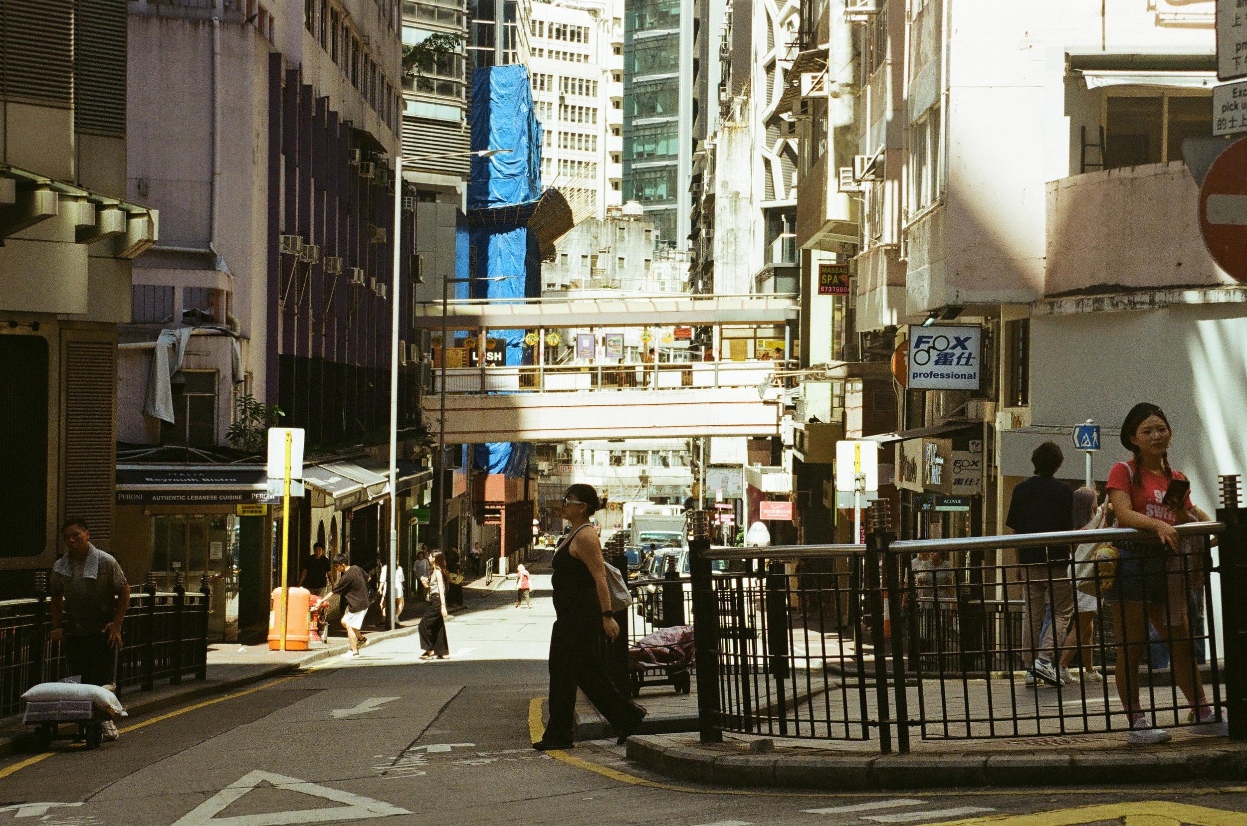 Urban street scene with pedestrians walking and standing on the sidewalk, multi-story buildings on both sides, and construction covered in blue tarp in the background.