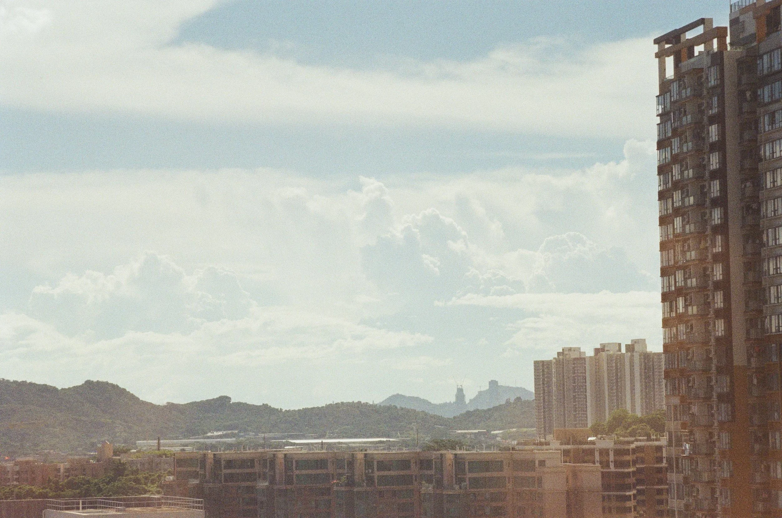 City skyline with high-rise apartment buildings, distant hills, and a partly cloudy sky.