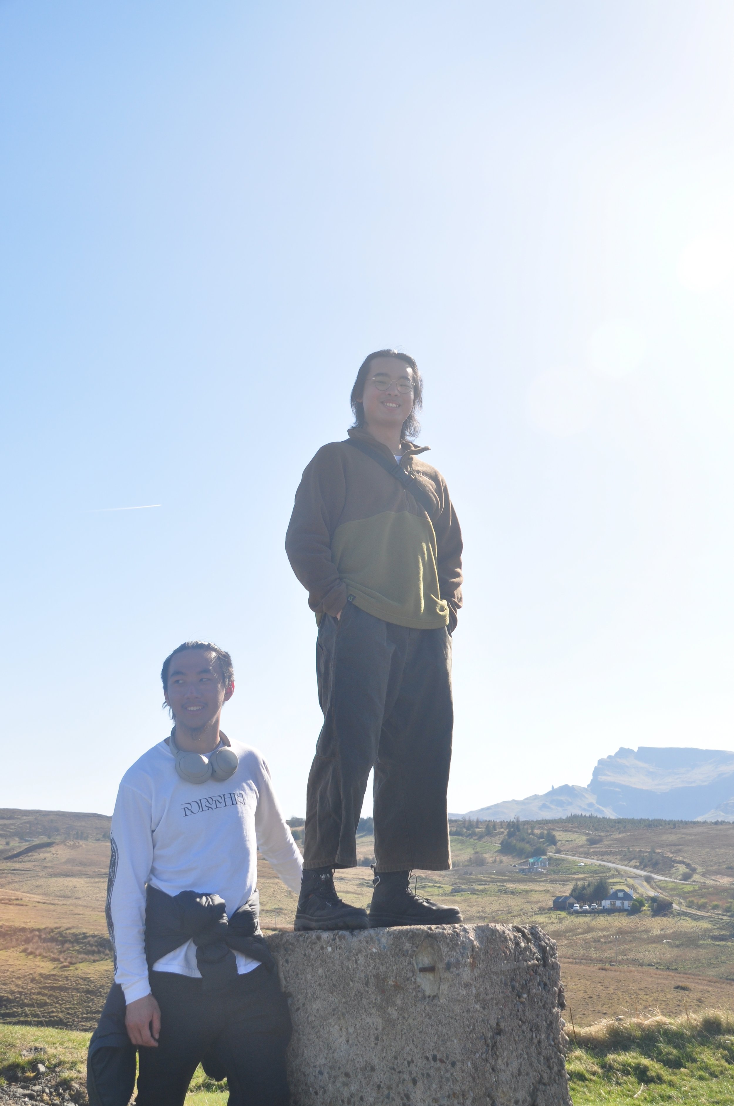 Two young adults in a scenic outdoor landscape, one standing on a large rock and the other standing beside it, under a clear blue sky with distant hills.