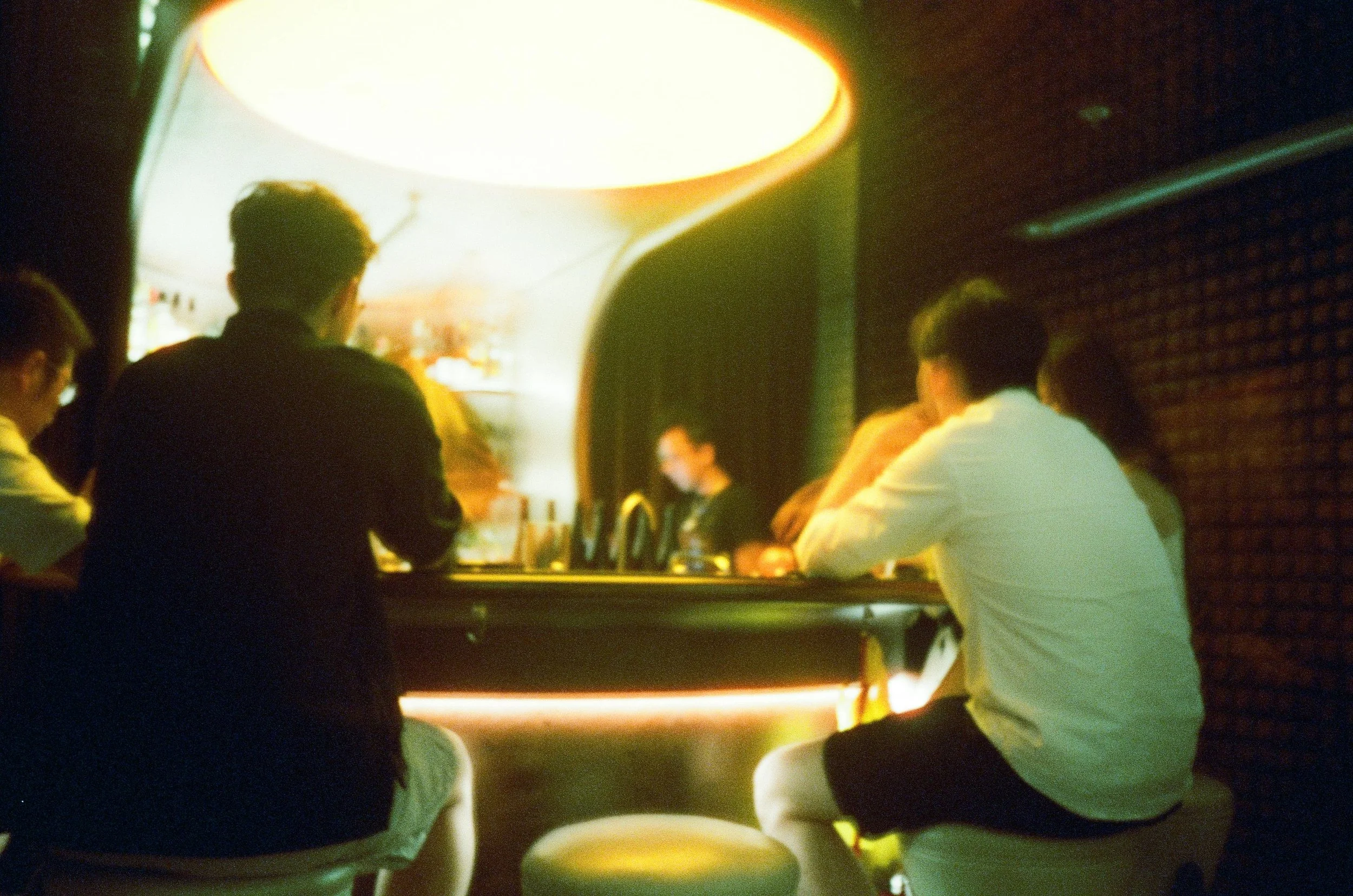 People sitting at a bar with a brightly lit open fridge behind them in a dimly lit setting.