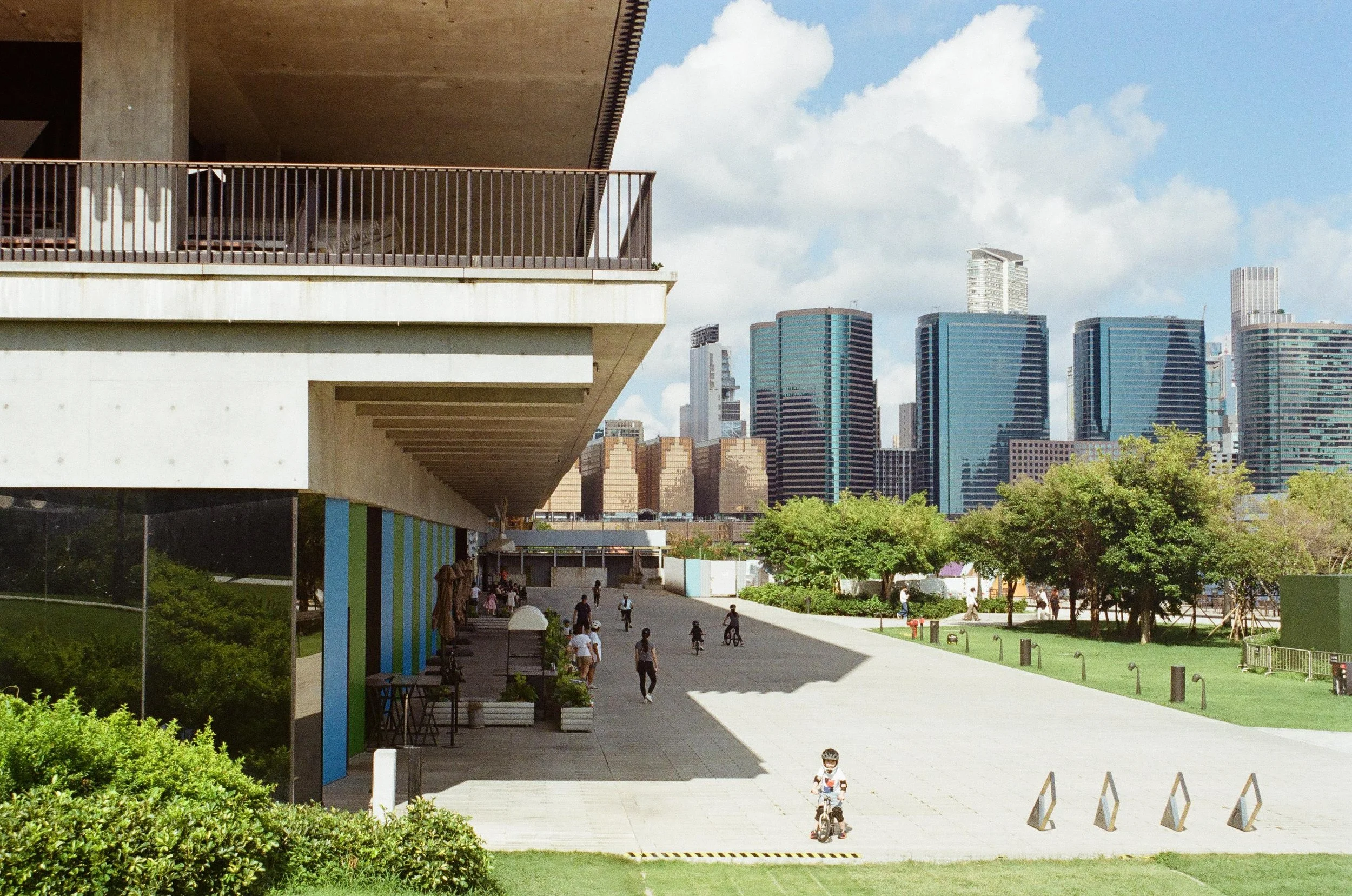 Urban park scene with people walking and riding bikes, modern buildings and skyscrapers in the background, trees and green grass, a concrete pathway, and a large building with an overhanging balcony.