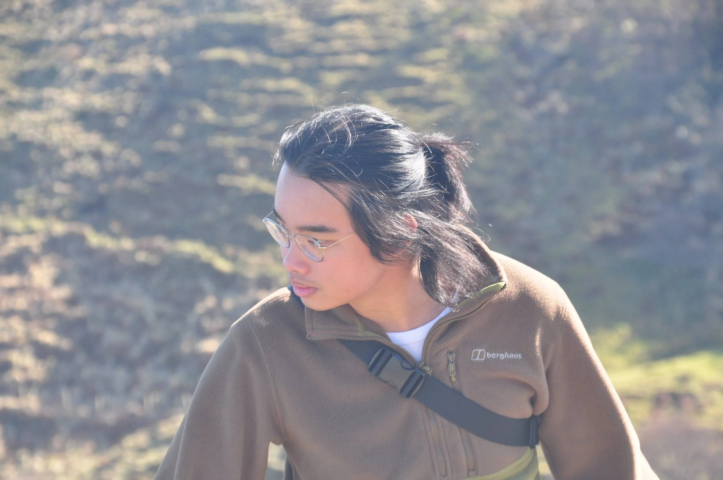 A young man with long dark hair, glasses, wearing a brown jacket with a zip, and a black strap across his chest, outdoors in a sunny, rocky landscape.