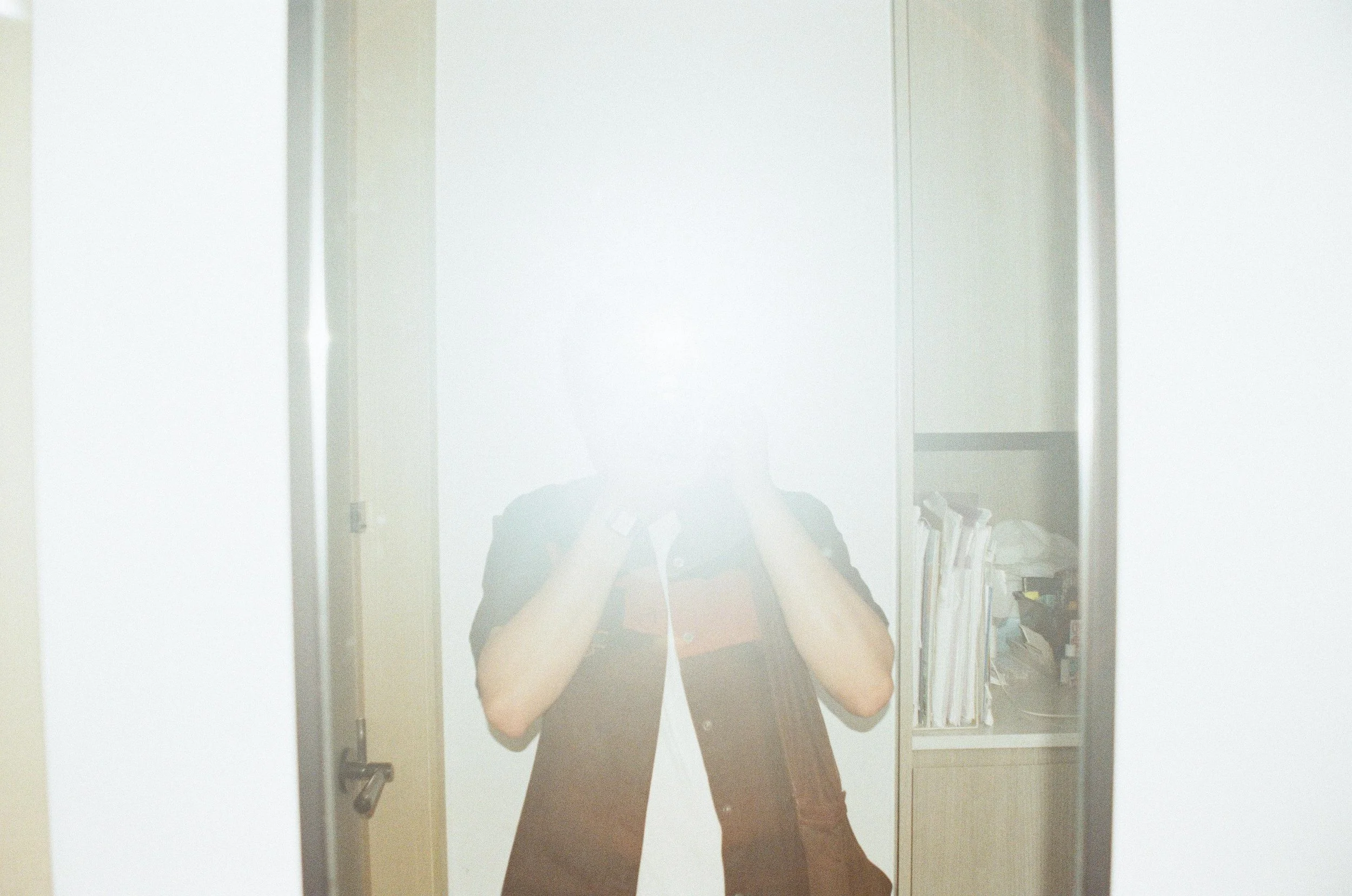Person taking a photo of themselves in a mirror, with their face obscured by the camera, in a room with a cabinet filled with documents and supplies.