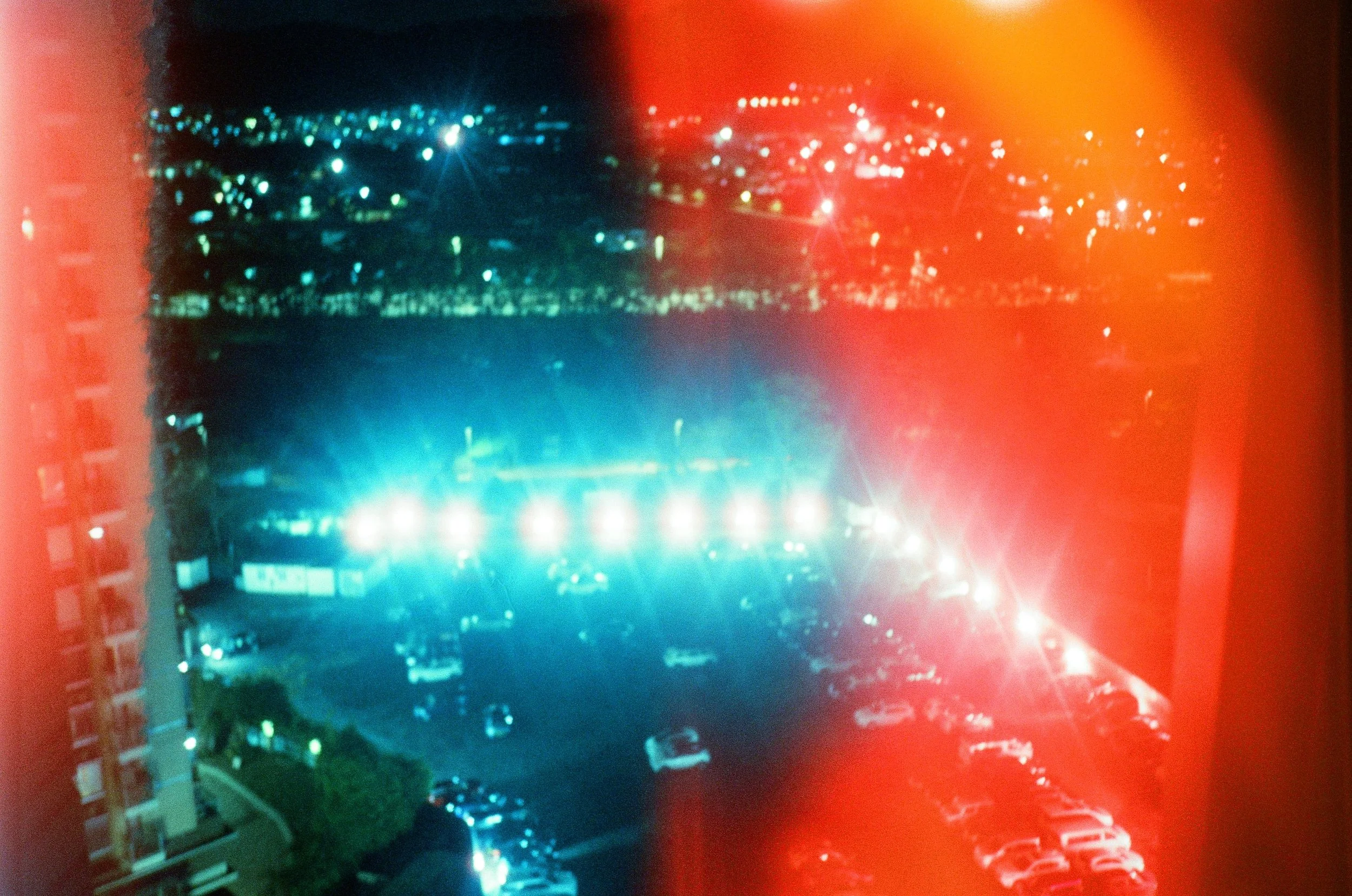 Nighttime view of a parking lot filled with cars, illuminated by streetlights and surrounded by a cityscape with buildings and more lights in the distance, framed by a red-orange light flare.
