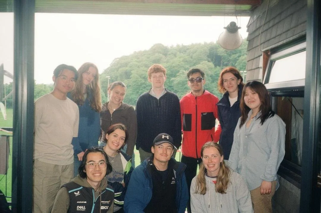 Group of 11 young adults standing and seated indoors, with a window showing trees and hills outside in the background.