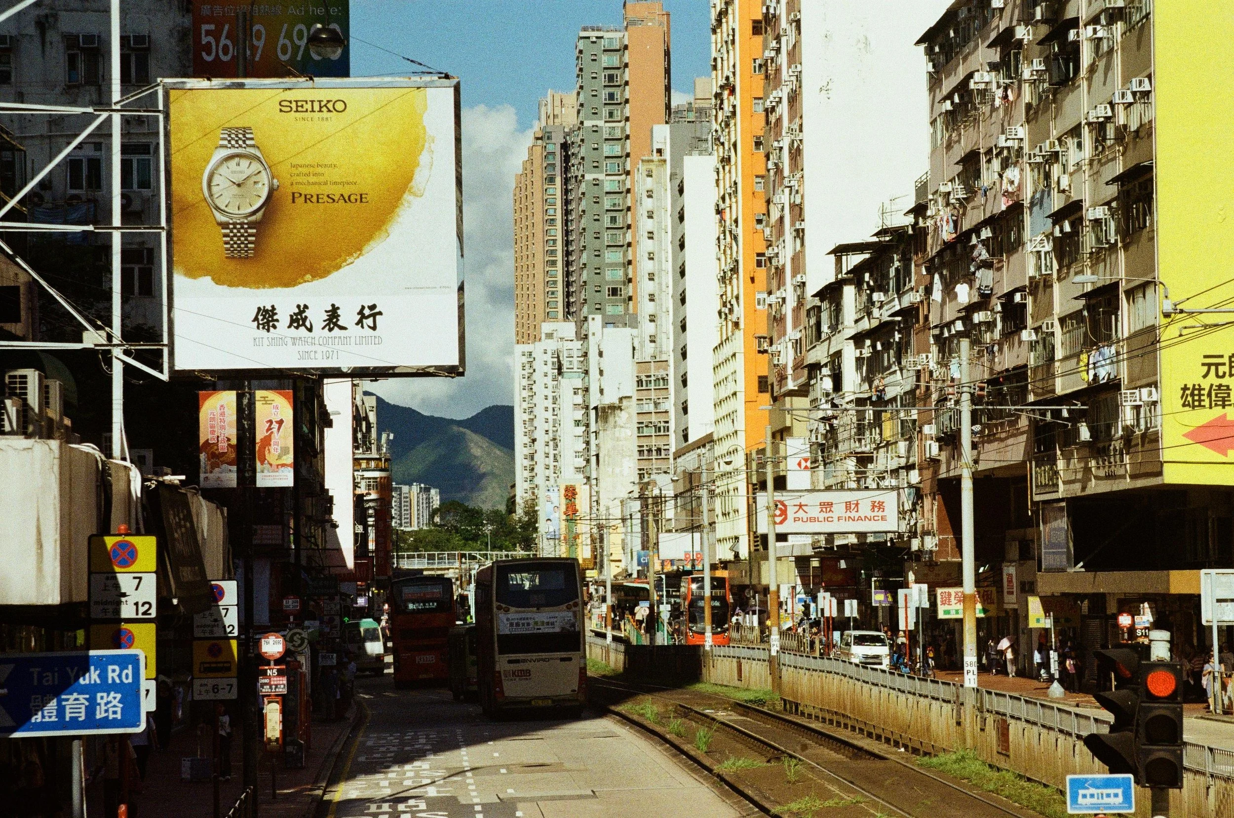 City street scene with tall residential and commercial buildings, buses and cars on the road, and various signs and advertisements, including a large yellow advertisement for Seiko watches with mountain in the background.