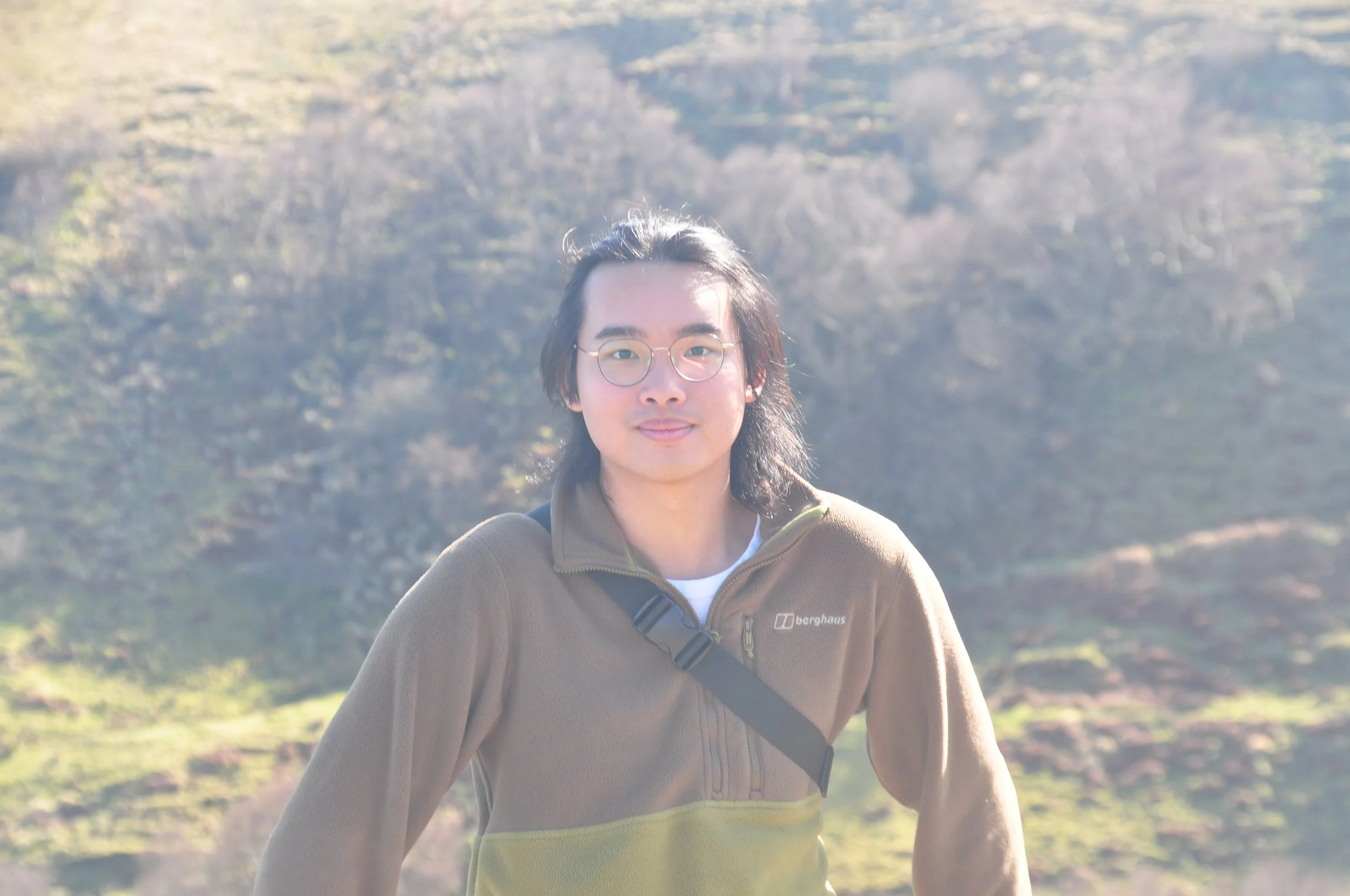 A young man with long dark hair and glasses outdoors in a hilly area with sparse vegetation, wearing a brown fleece jacket and a black strap across his chest.