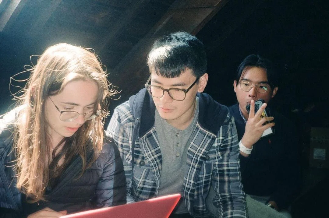 Three young adults, two men and one woman, wearing glasses, look at a red device in a dimly lit setting, possibly a bar or restaurant.
