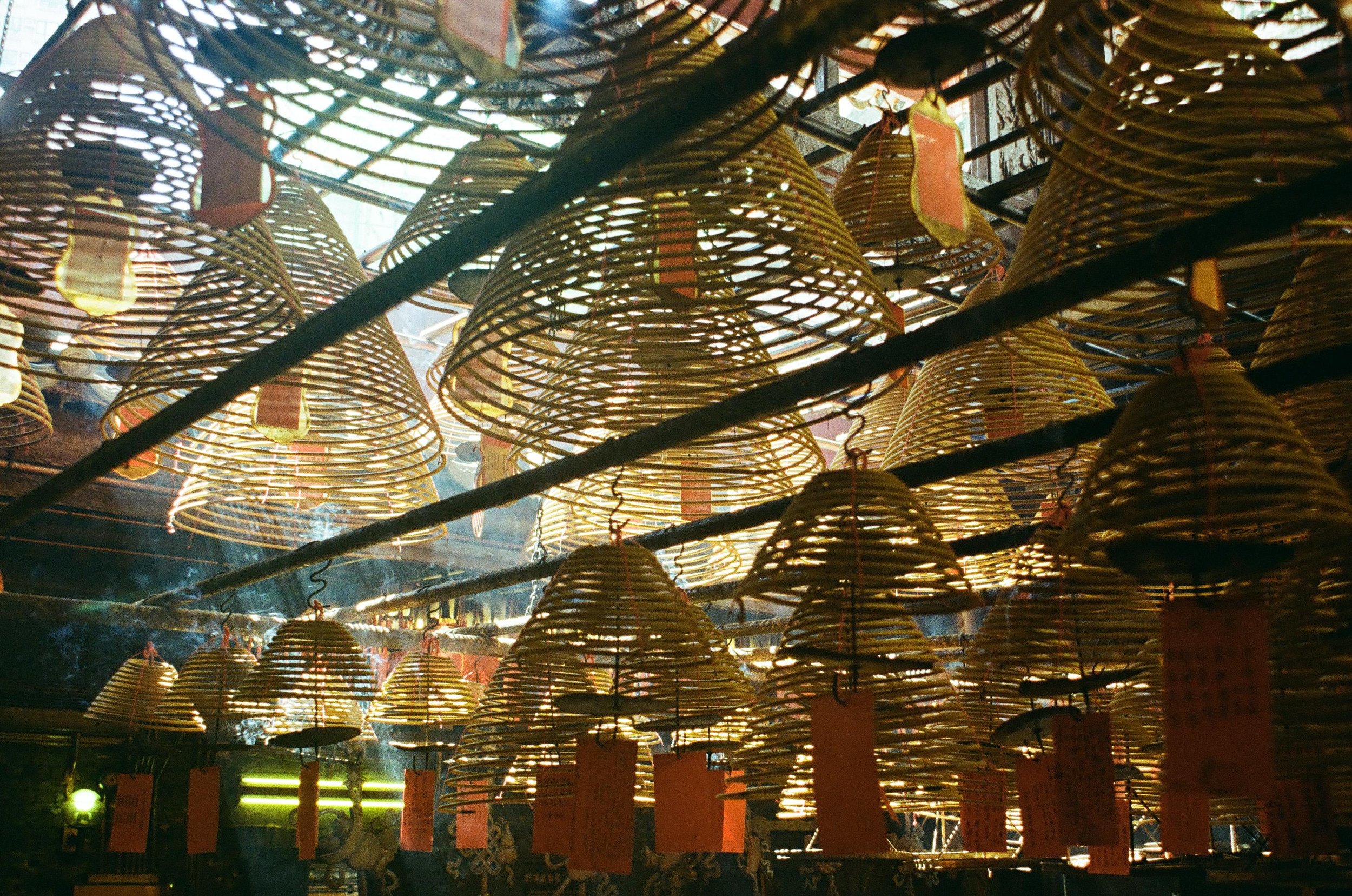 Multiple handmade bamboo prayer lanterns hanging in a temple, with some smoke and sunlight filtering through.