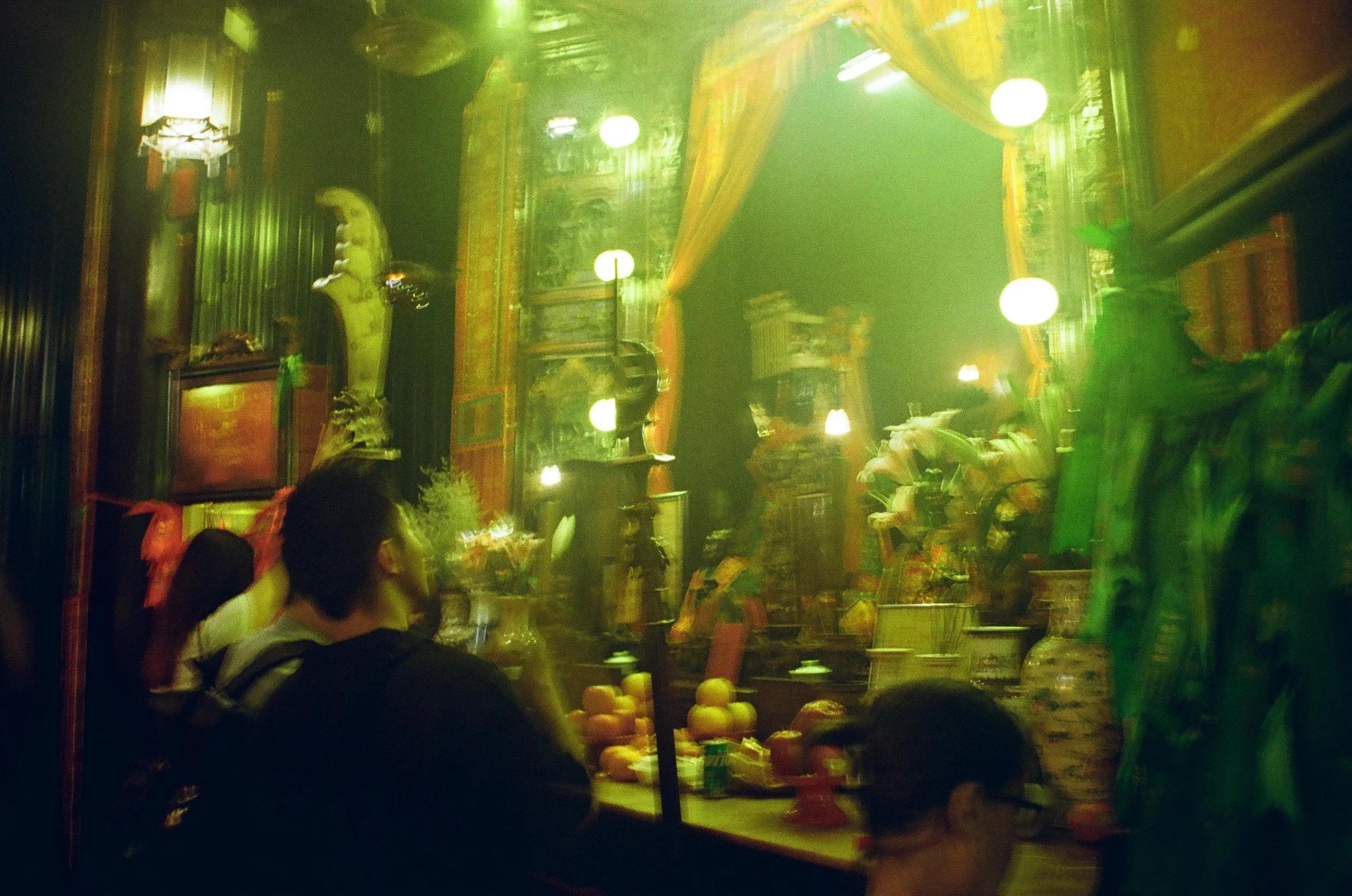 A dimly lit, ornate setting resembling a temple or shrine with traditional Asian decor. There are flowers, fruit, and candles on display, along with statues and paintings. Several people are seated and observing the surroundings.