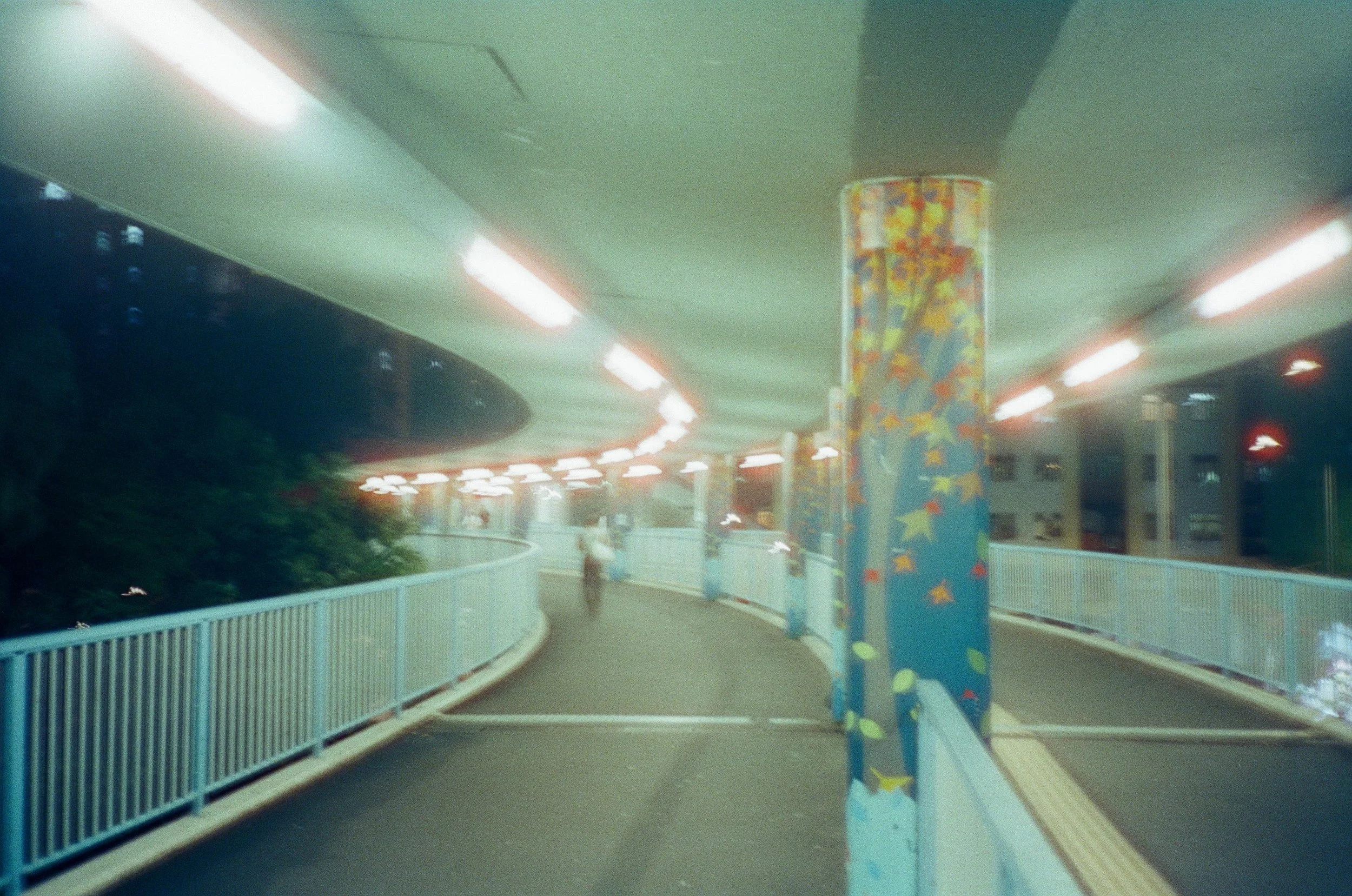 An indoor pedestrian walkway with a curved path, a blue pillar decorated with colorful shapes, bright ceiling lights, a white railing on the sides, and a few blurred people walking.