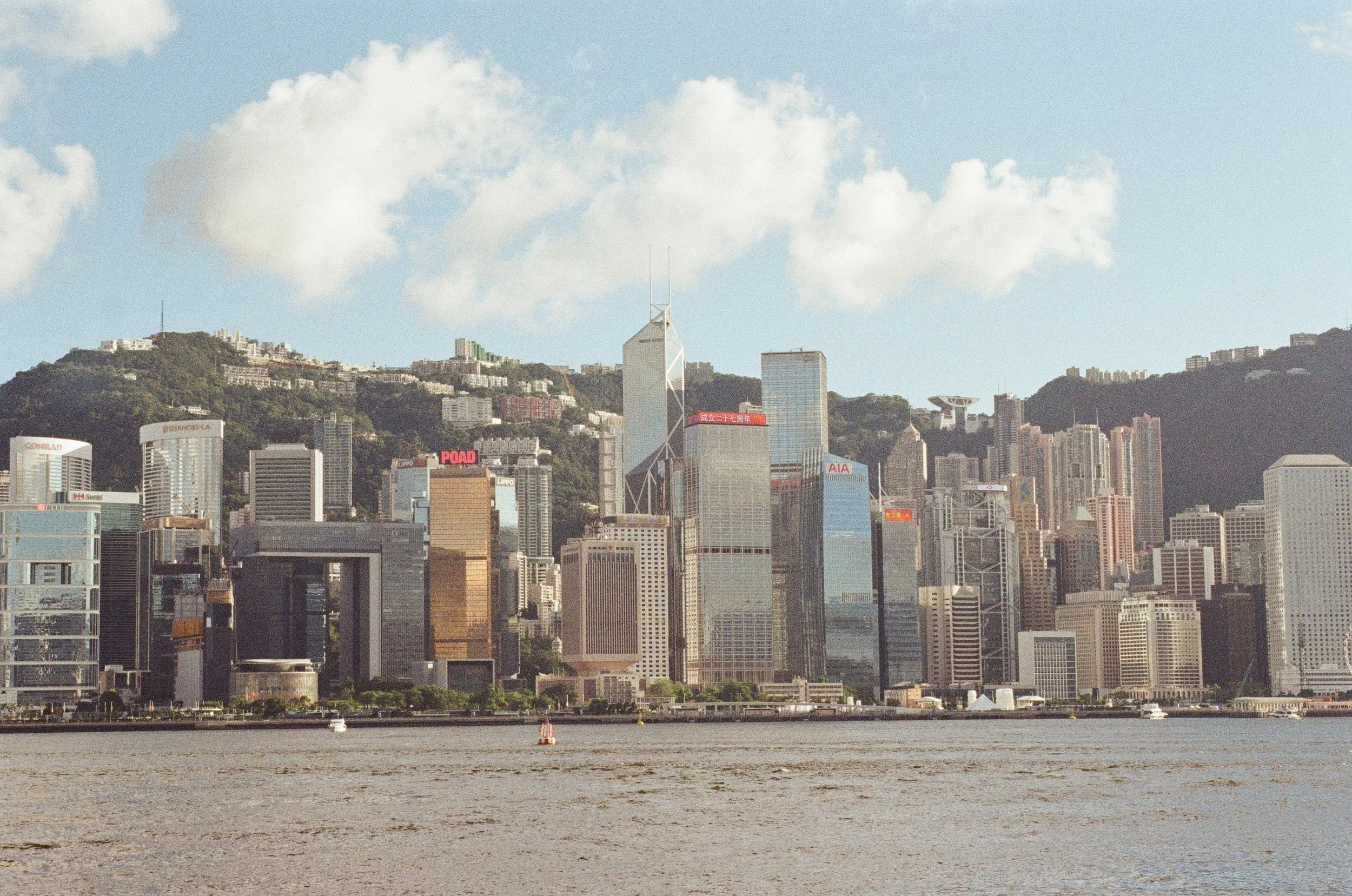 Hong Kong skyline with high-rise buildings and Victoria Harbour in the foreground, hills in the background, partly cloudy sky.