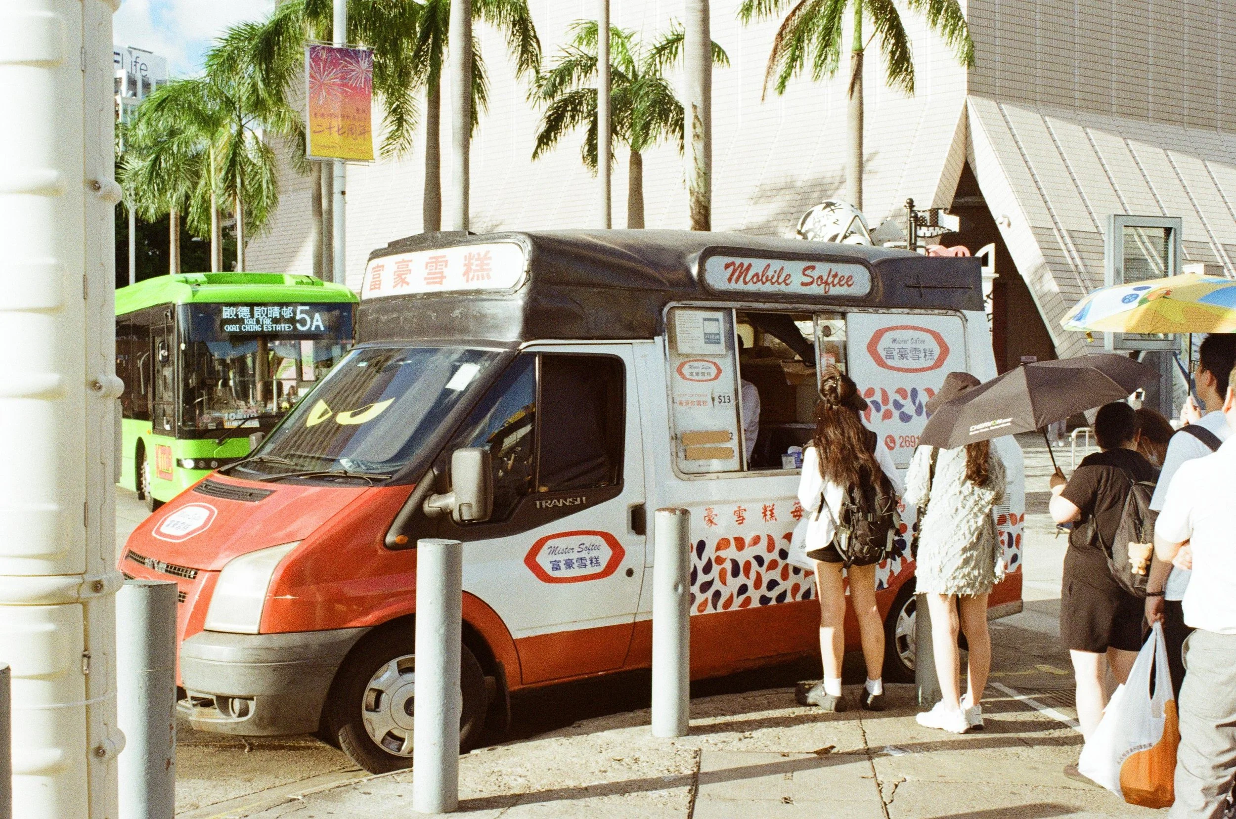 A red and white mobile ice cream truck with the words "Mobile Softee" on top, parked on a city street. Several people are standing around, some with umbrellas to shield from the sun, and a green bus in the background. Palm trees line the street.