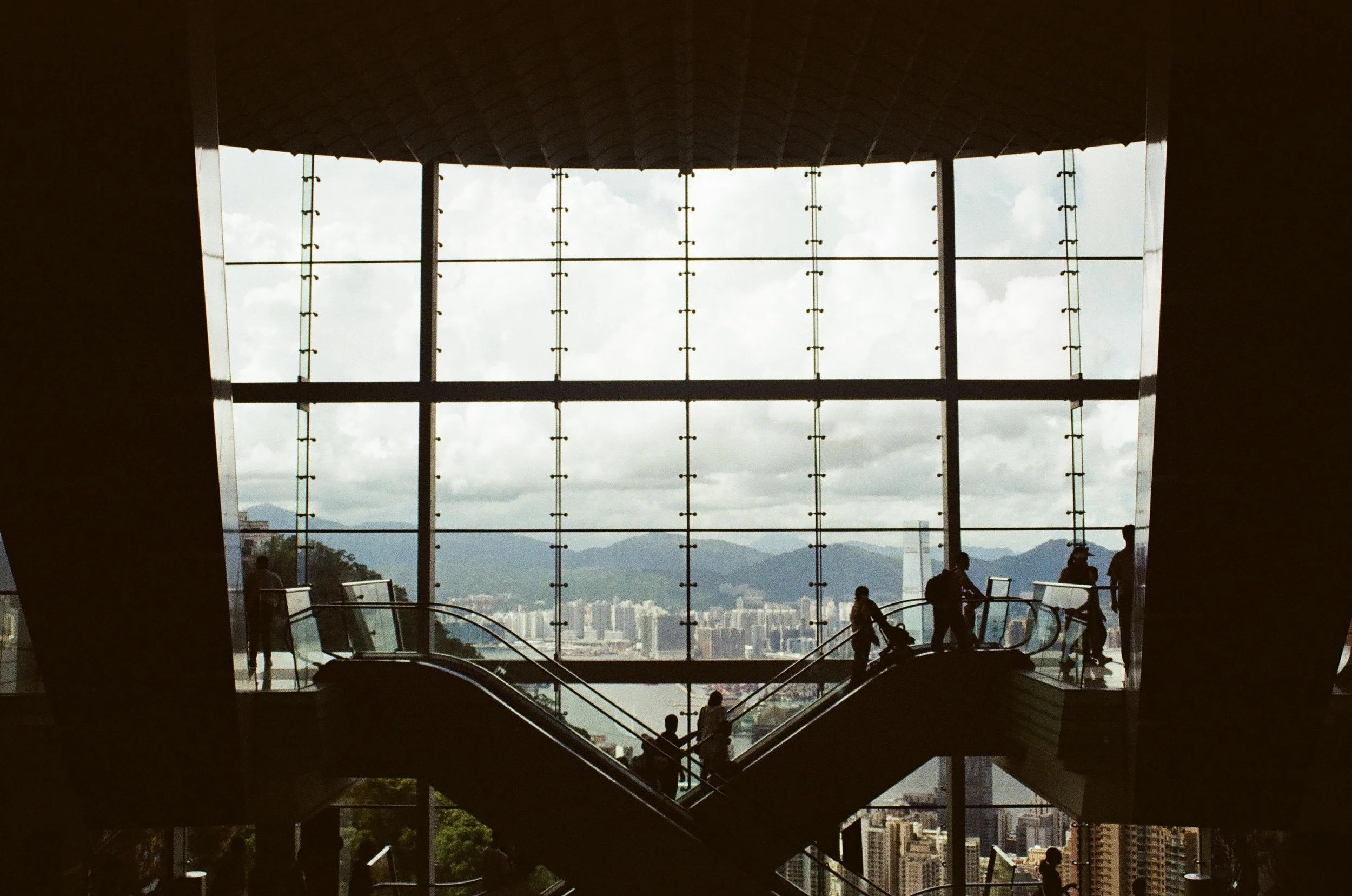 Interior of a modern building with large glass window panels offering a city and mountain view, people walking on escalators and viewing the scenery.