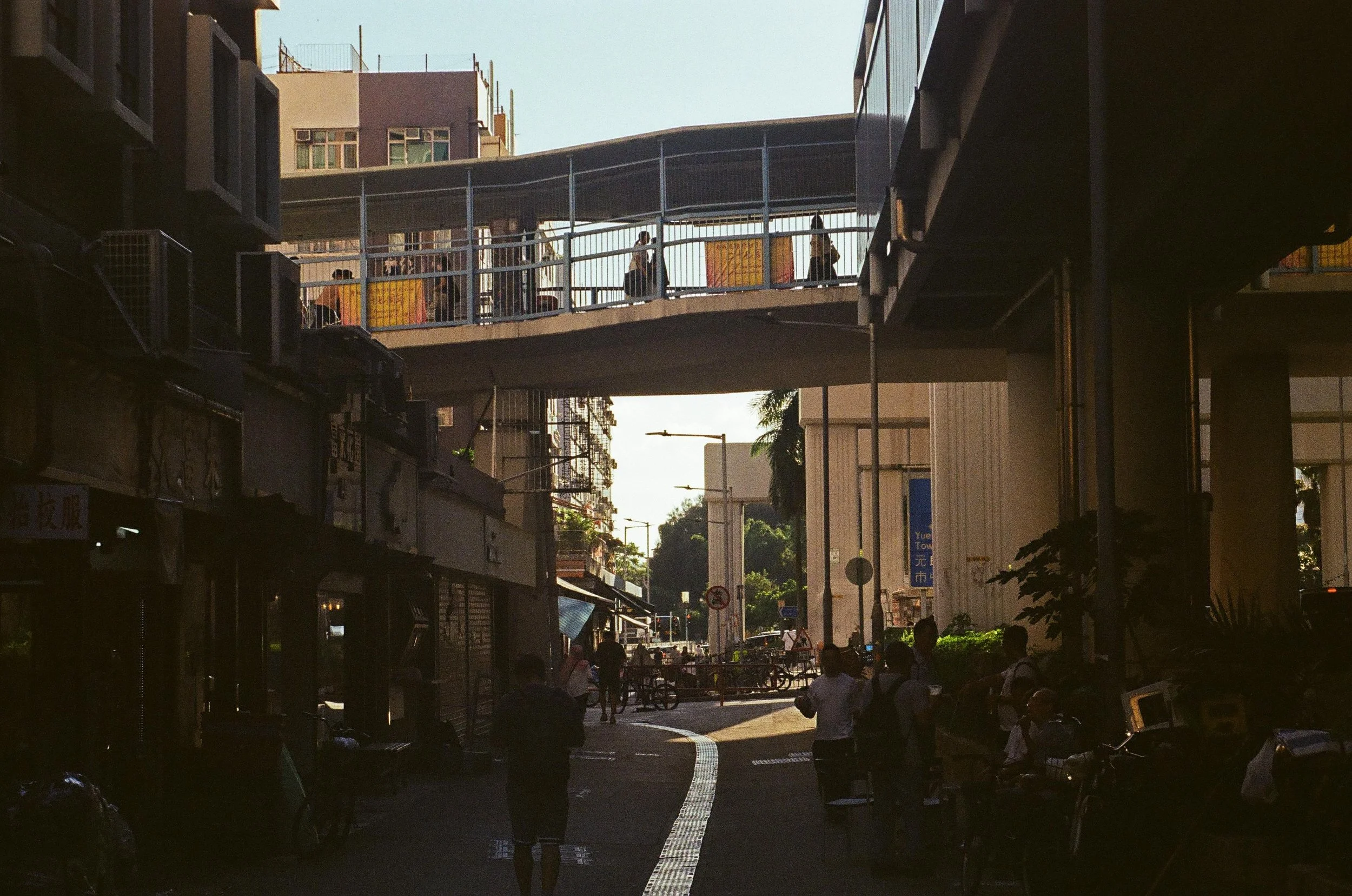 Urban street scene with pedestrians walking under elevated walkways, buildings, and trees in the background during daylight.
