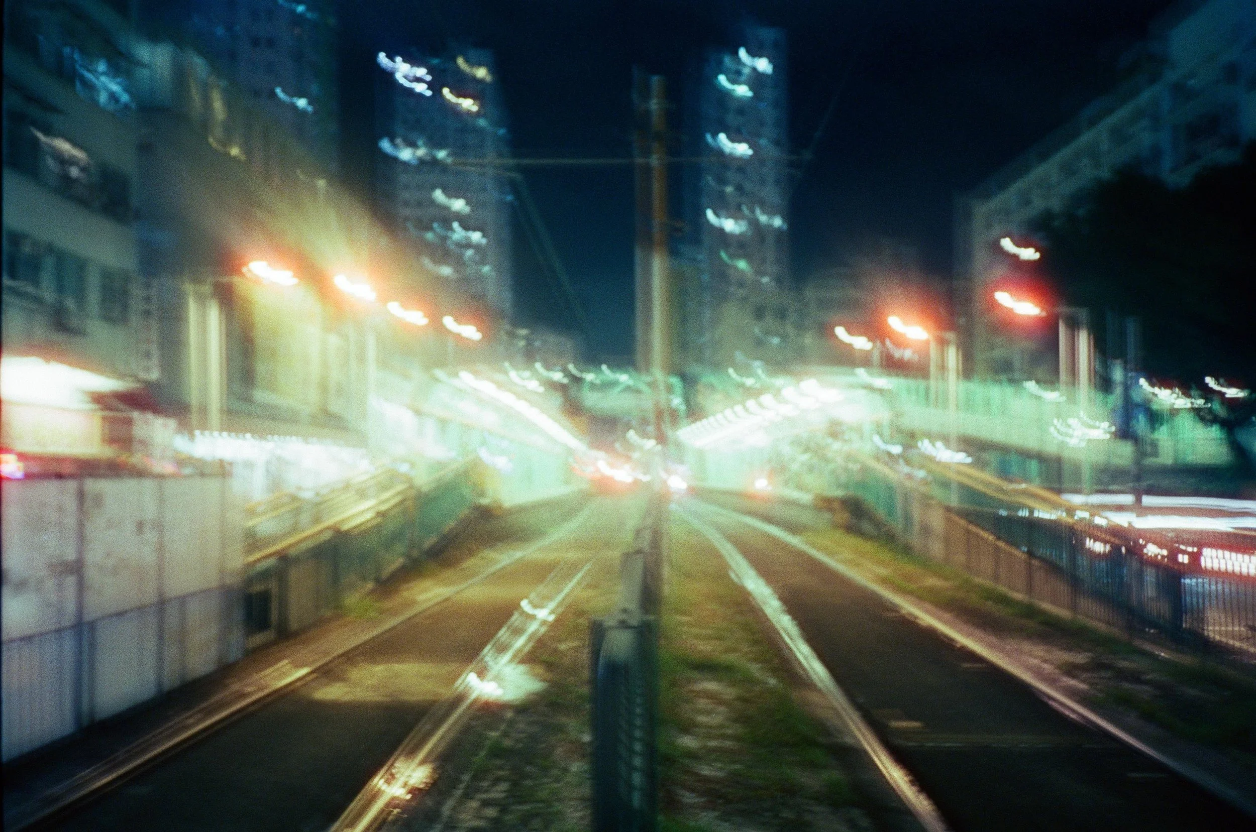 Blurry photo of a city street at night with train tracks, buildings, and streetlights.