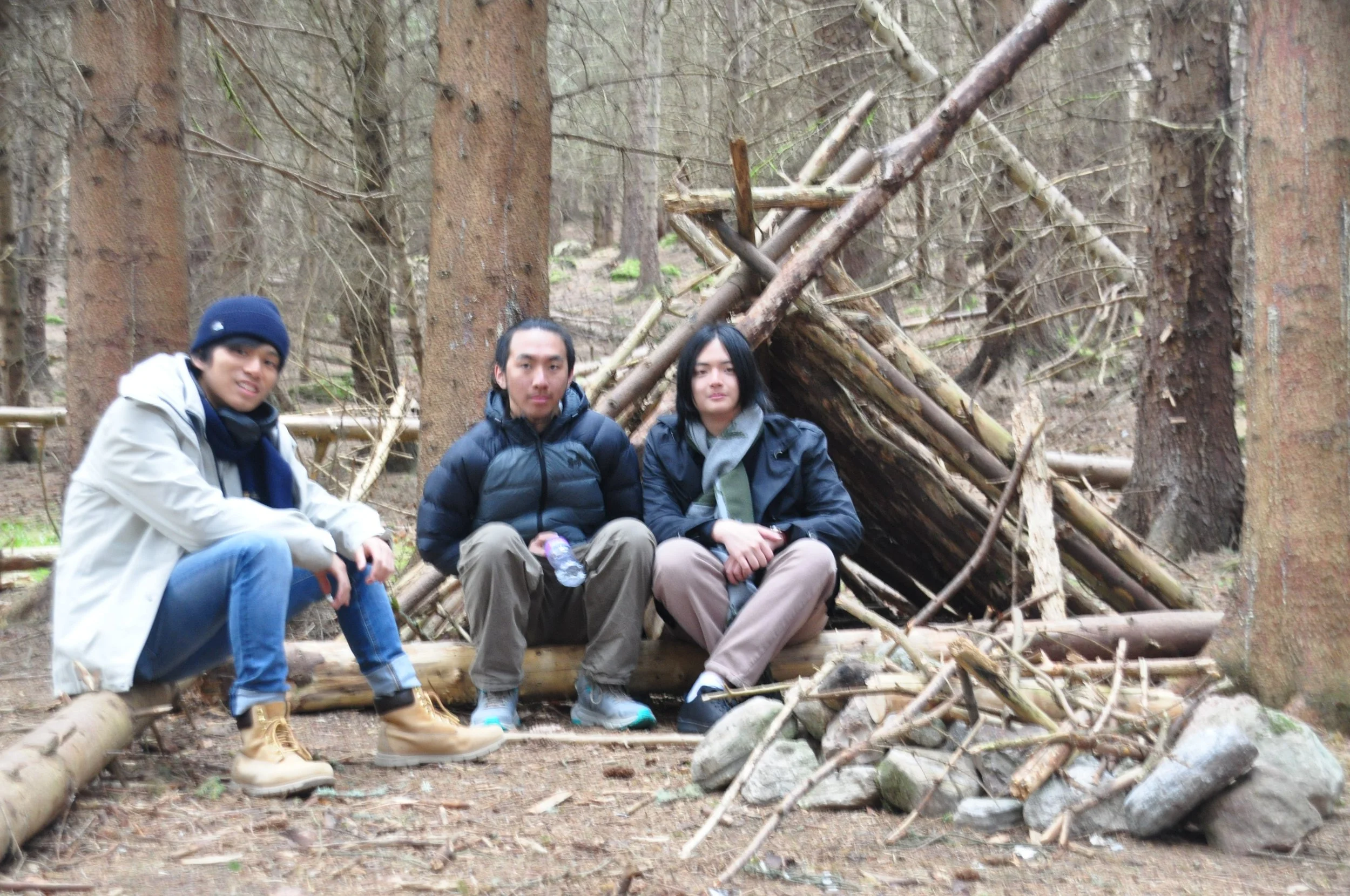 Three young men sitting on logs and rocks in a forest with a makeshift shelter made of sticks behind them.