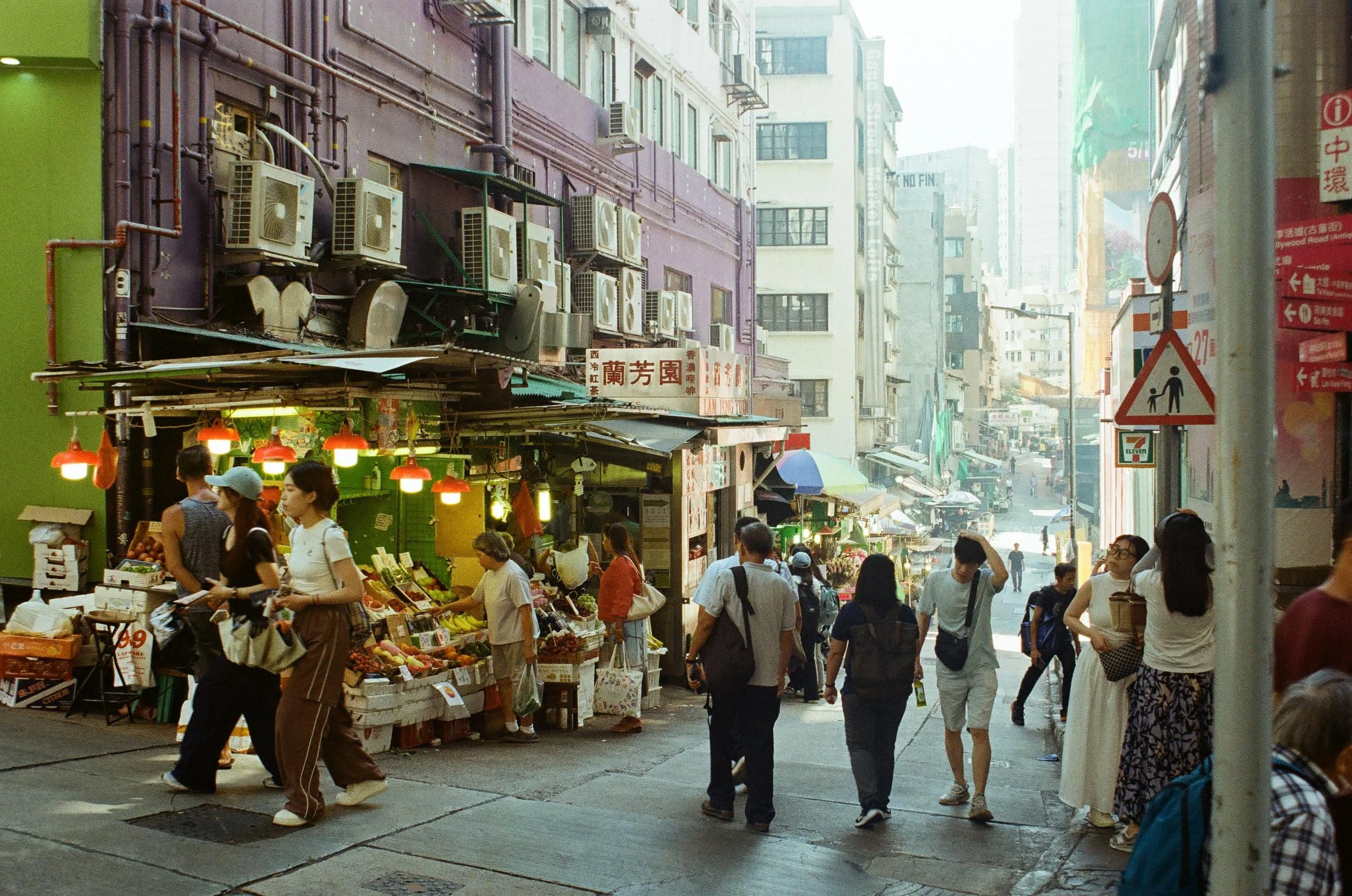 Street scene with people shopping at a fruit stall in an Asian city. Many pedestrians walk along the sidewalk with high-rise buildings in the background.