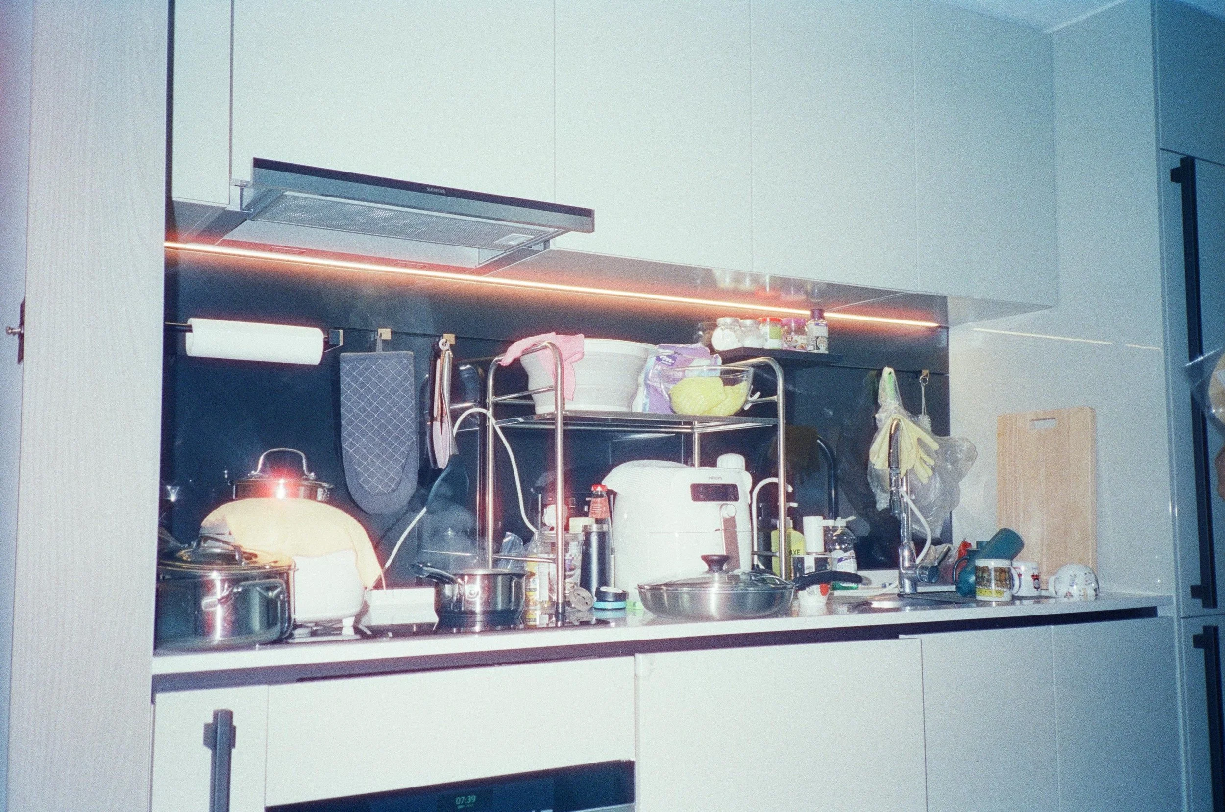 A cluttered kitchen countertop with various kitchen appliances, utensils, and containers, including a rice cooker, pots, and a dish rack, against a backdrop of modern cabinetry and a stainless steel backsplash.
