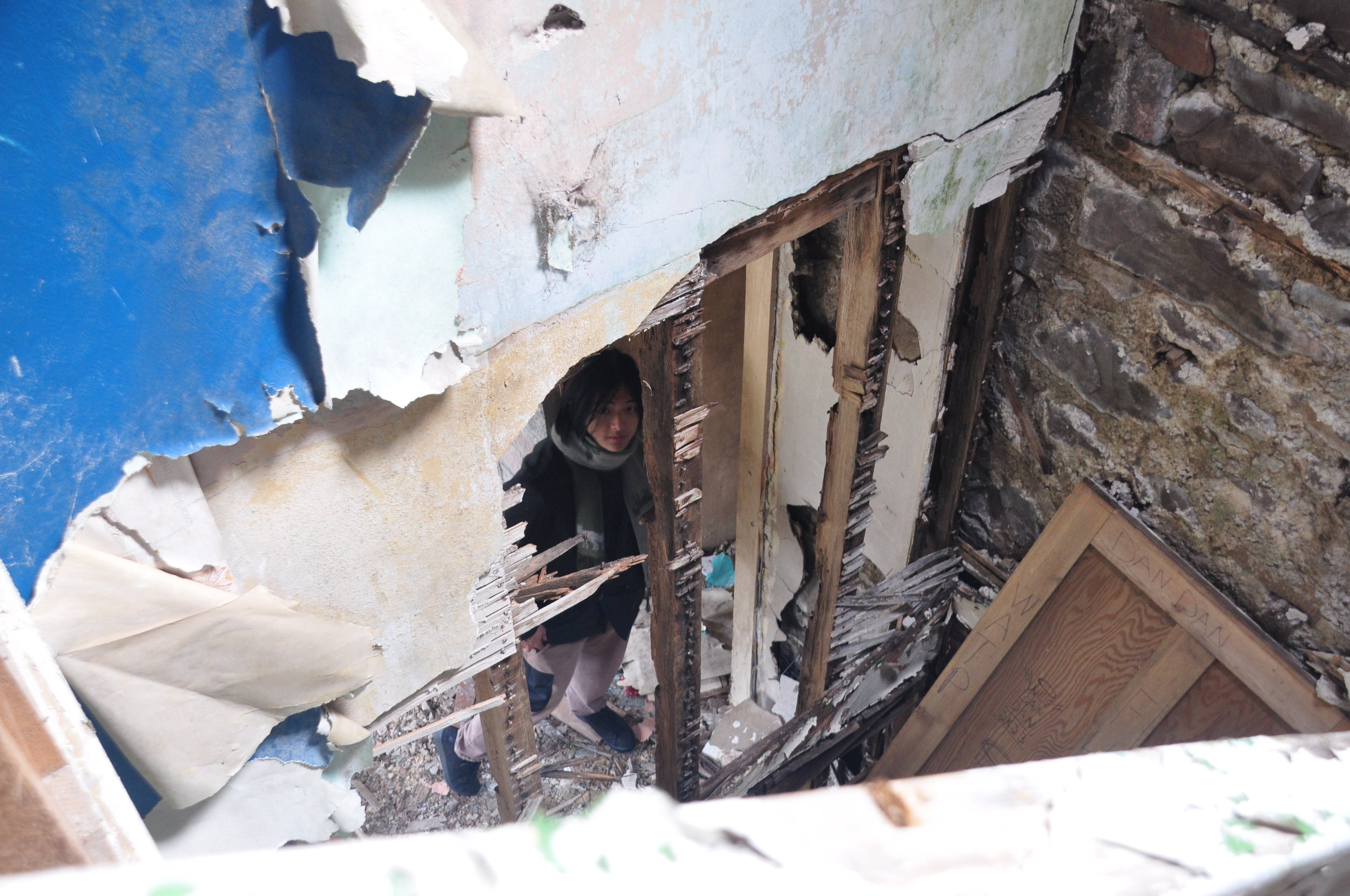 A woman standing in a partially demolished room with exposed wooden studs and damaged walls, looking up through a hole in the ceiling.
