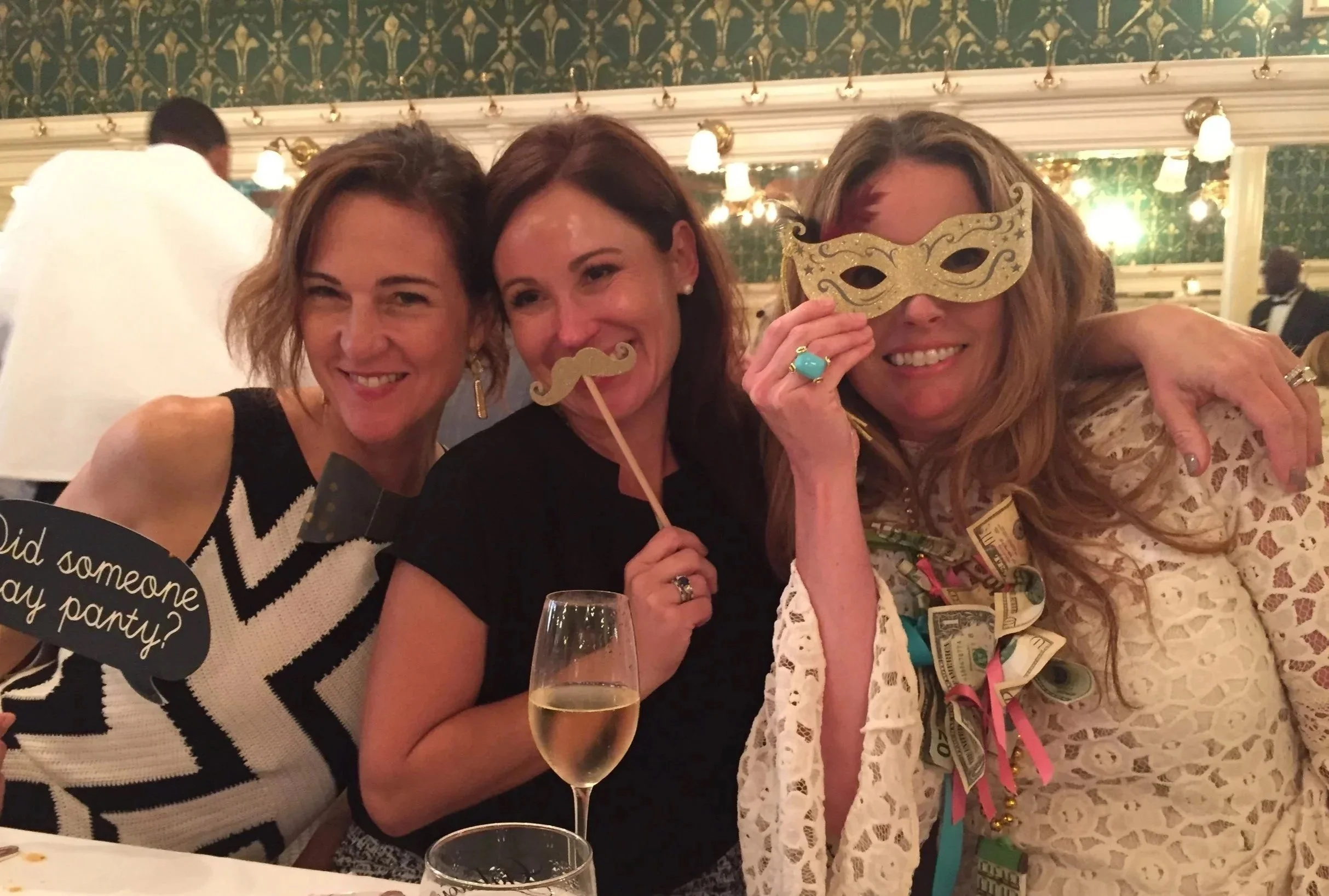 Three women at a celebration, smiling, with one woman holding a gold masquerade mask, another holding a stick with a gold mustache, and the third holding a black speech bubble prop that says 'Did someone say party?'. One woman has dollar bills around her neck, and there are drinks on the table in front of them.