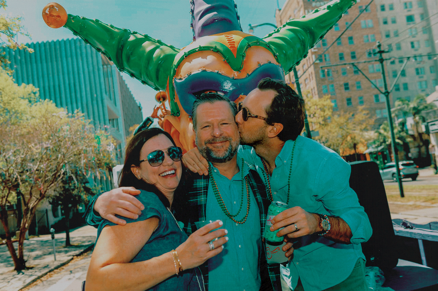 Three adults celebrating outdoors with a large jester float in the background during a sunny day. One man is kissing another man on the cheek, both are smiling, and a woman with sunglasses is smiling and holding a drink. The scene is on a city street with trees and buildings.