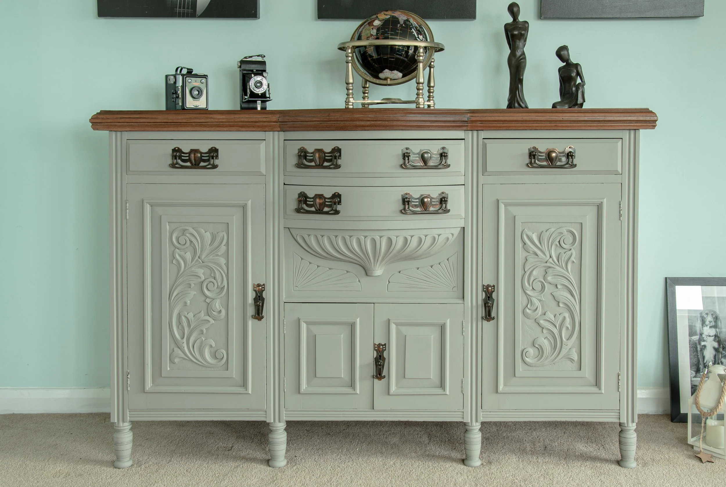 A antique sideboard with intricate carvings and a wooden top, placed against a light green wall. Decorative items on top include vintage cameras, a globe, and abstract human figurines. A framed photograph is on the floor leaning against the wall on the right side.