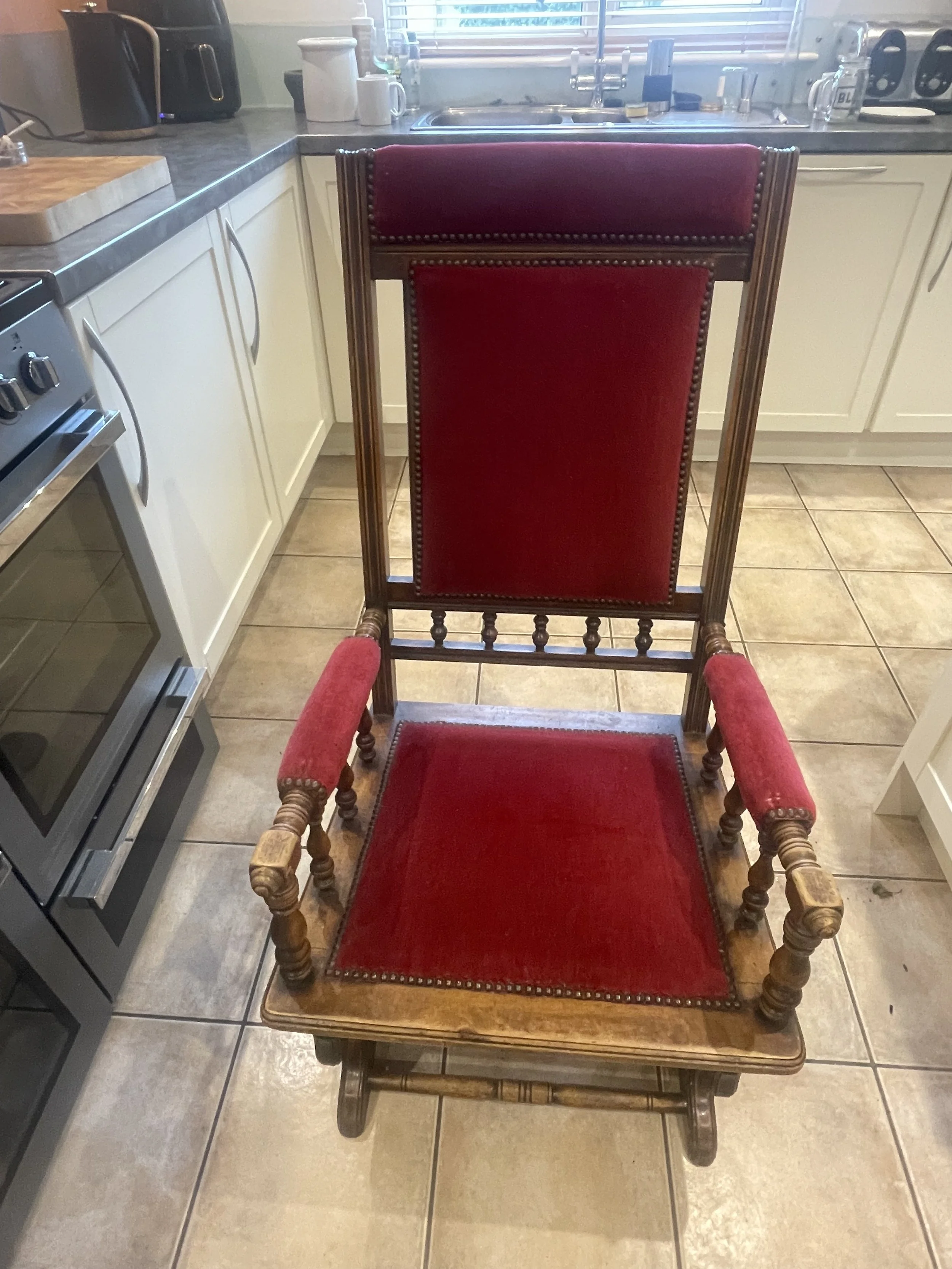 A wooden rocking chair with red velvet upholstery on the backrest and seat, in a kitchen with white cabinets, a gray countertop, and tiled floor.