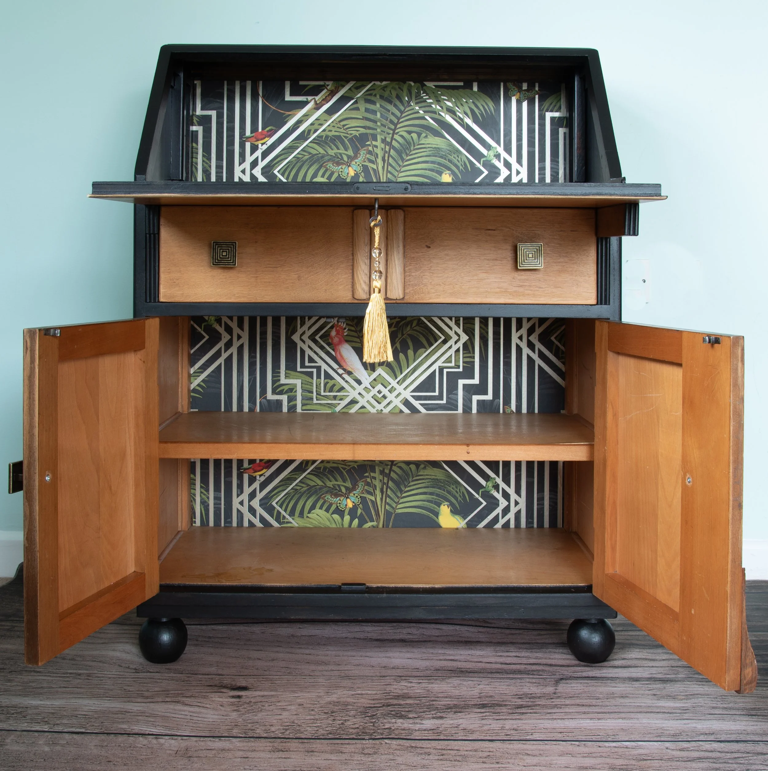 An open wooden cabinet with a decorative back panel, featuring a tropical foliage and butterfly pattern, and a small drawer with a tassel pull at the top.