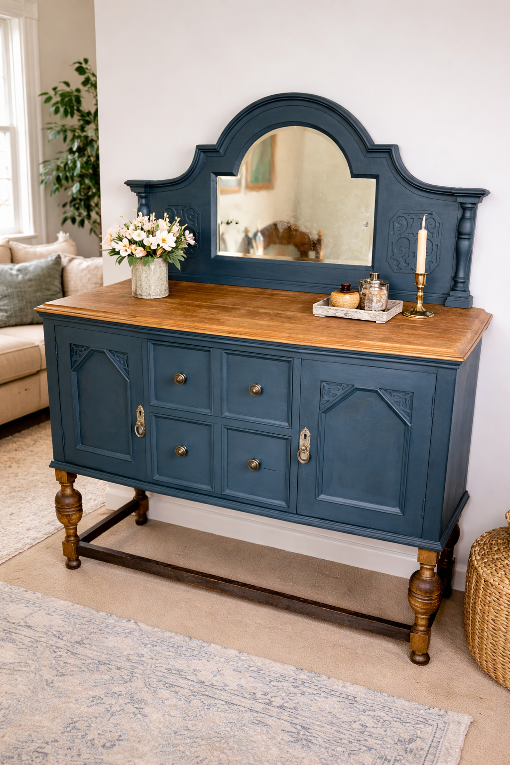 A vintage blue wooden sideboard with a large mirror on top, decorated with a vase of white and pink flowers, a candle, and small decorative containers, in a cozy living room.
