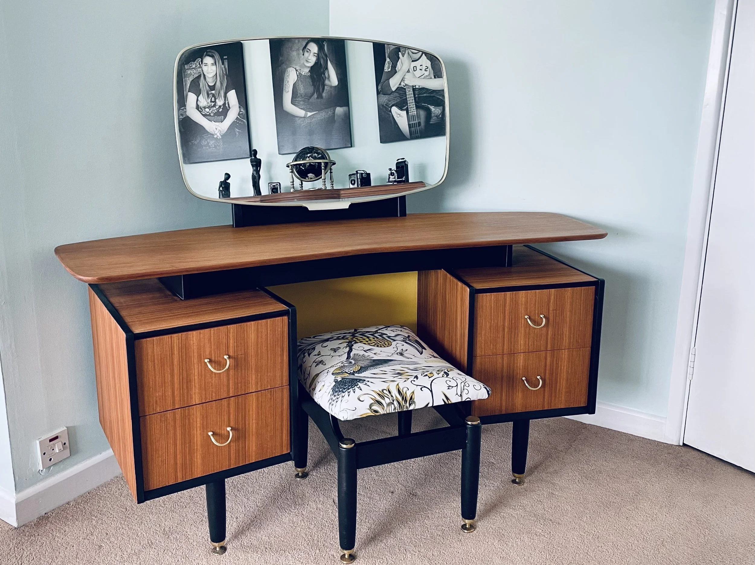 Vintage wooden vanity desk with black metal accents, a matching stool with a patterned cushion, a mirror with three framed black-and-white photographs, and various decorative items on the top shelf, against a light green wall.