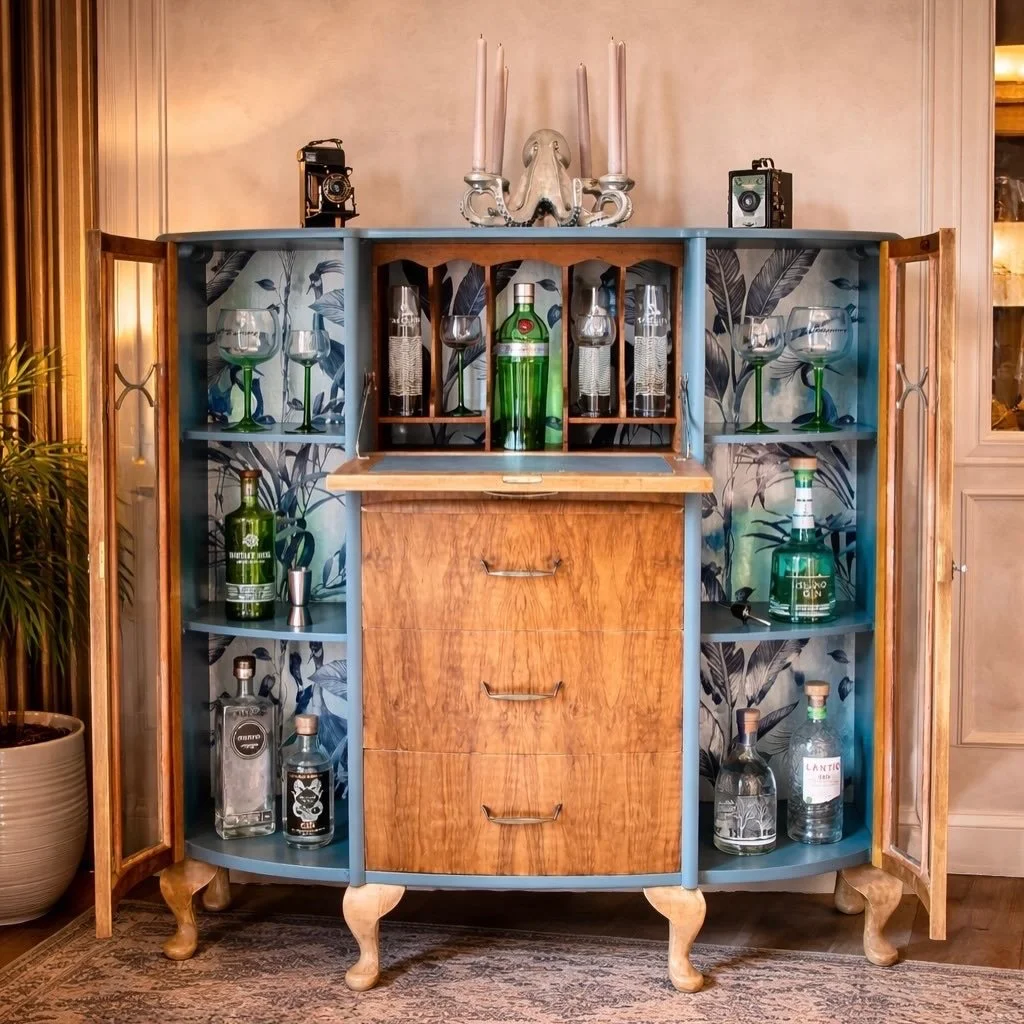 A vintage-style bar cabinet with open doors revealing bottles of gin and glasses inside. The cabinet has a wooden front with three drawers, blue sides and framing, and botanical wallpaper inside. On top of the cabinet, there are decorative items including a flamingo-shaped holder with candles, two old-fashioned cameras, and a large green bottle of Tanqueray gin.