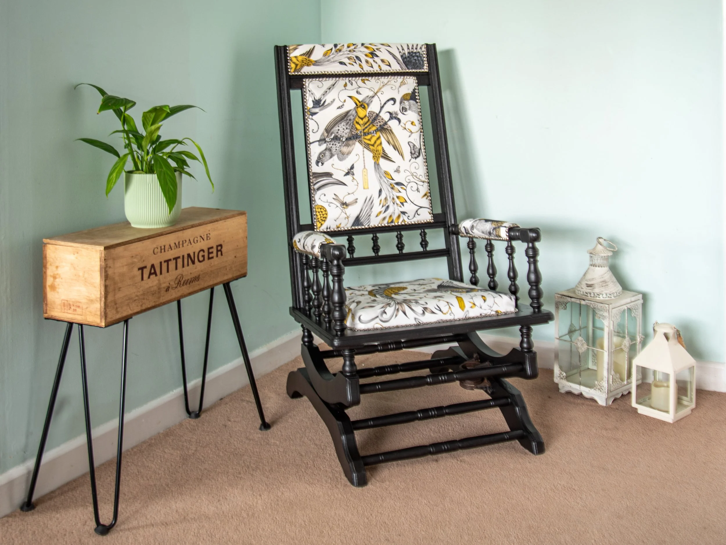 A black wooden rocking chair with floral-patterned cushions featuring yellow, black, and white colors, positioned in a corner of a room with a light wall. To the left, there is a small wooden stand with a potted green plant. To the right, there are two white lanterns, one with a handle and candle inside, placed on the floor.