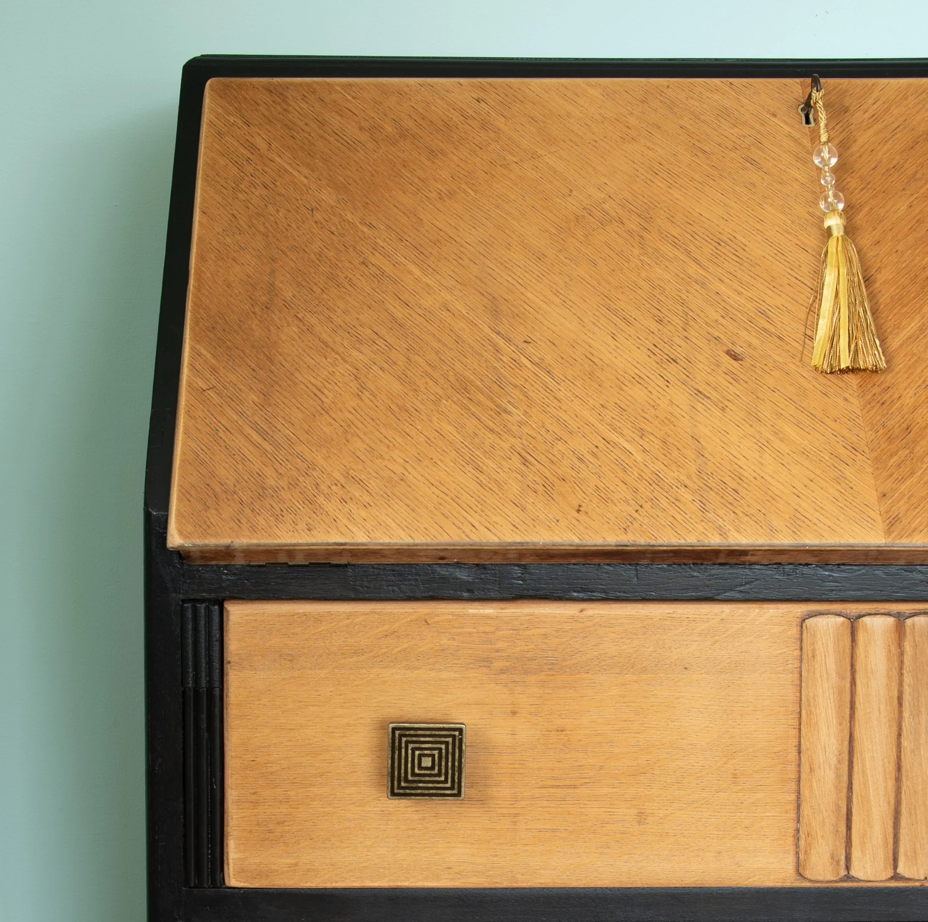 Close-up of a vintage wooden desk or cabinet with a single drawer featuring a square hardware handle, and a decorative tassel hanging from the corner of the wooden top, set against a mint green wall.