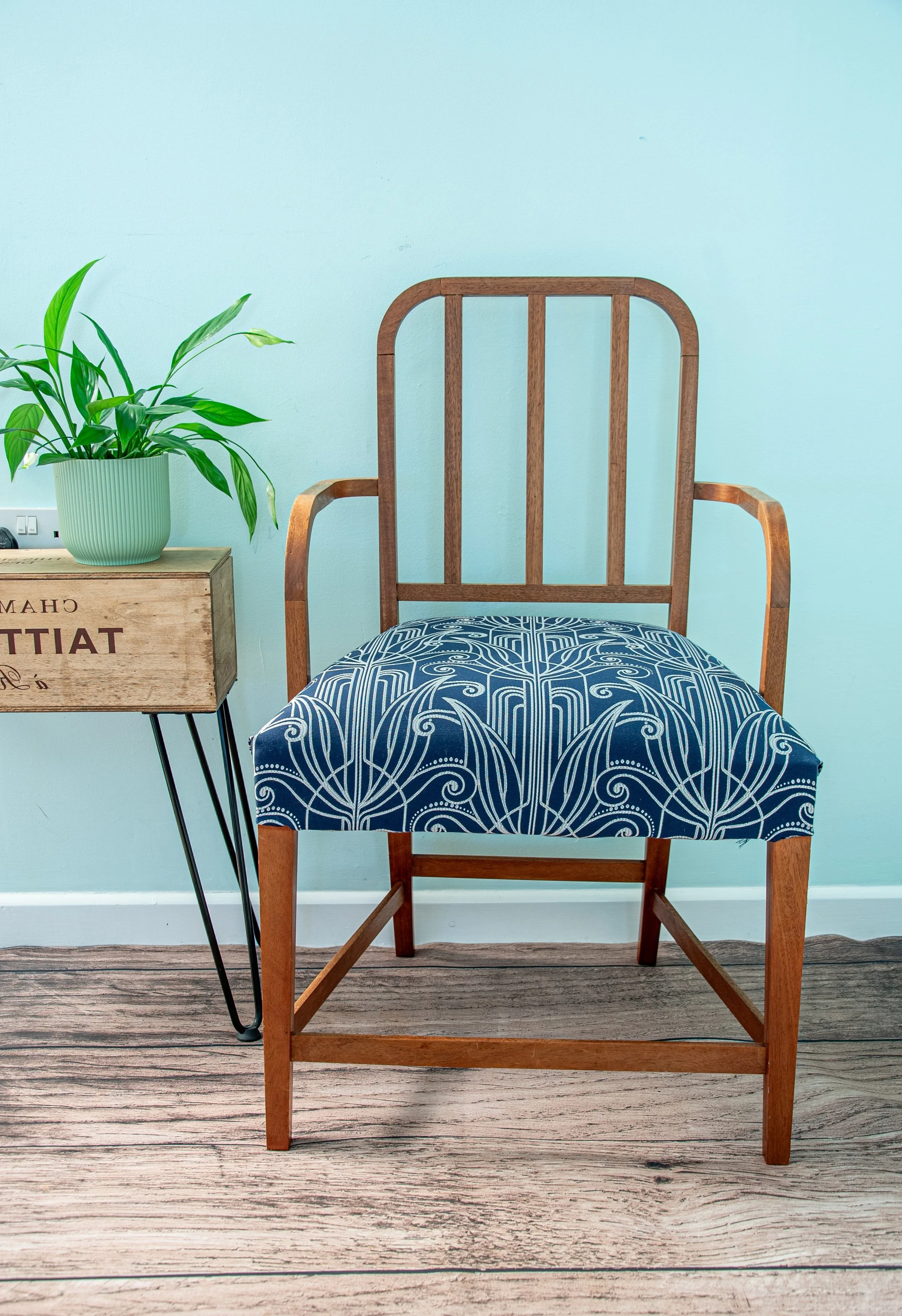 A wooden chair with a decorative blue and white cushion, placed next to a small wooden side table with a potted plant on top, against a light blue wall.