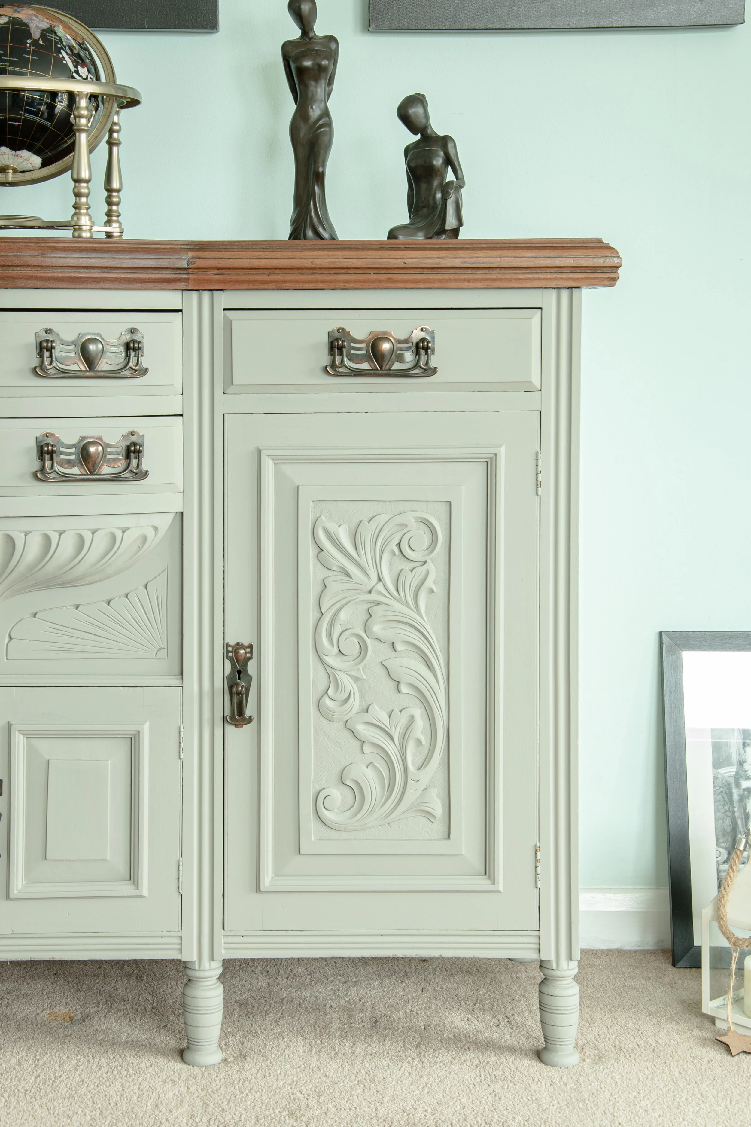 Decorative sideboard with ornate carvings, topped with two bronze statues of women, an international globe, and a framed picture resting against the wall.