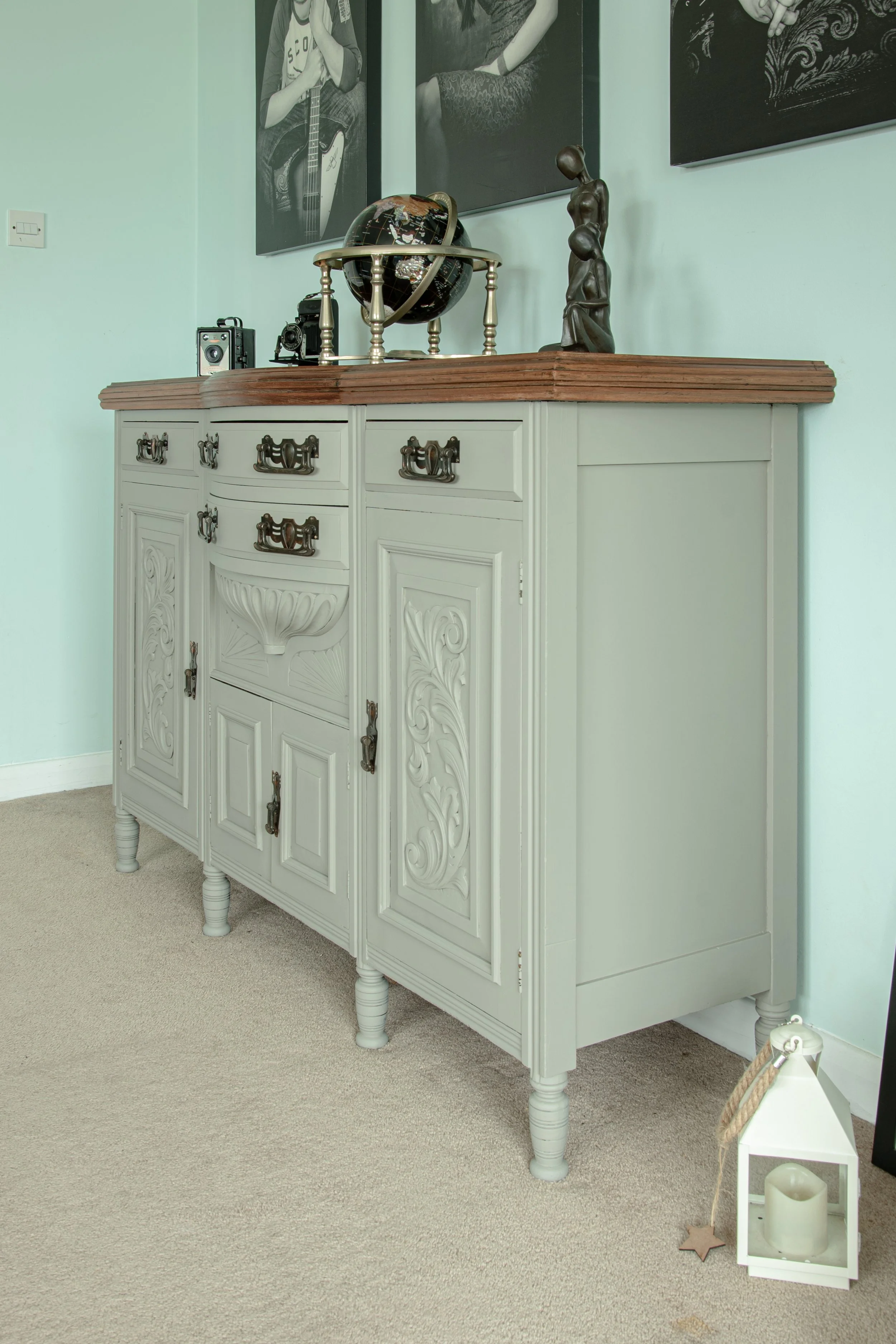 Decorative sideboard with ornate detailing and brass handles, topped with a vintage globe, a small camera, and a sculpture of a woman in a dress, set against a light green wall with black and white framed photographs.