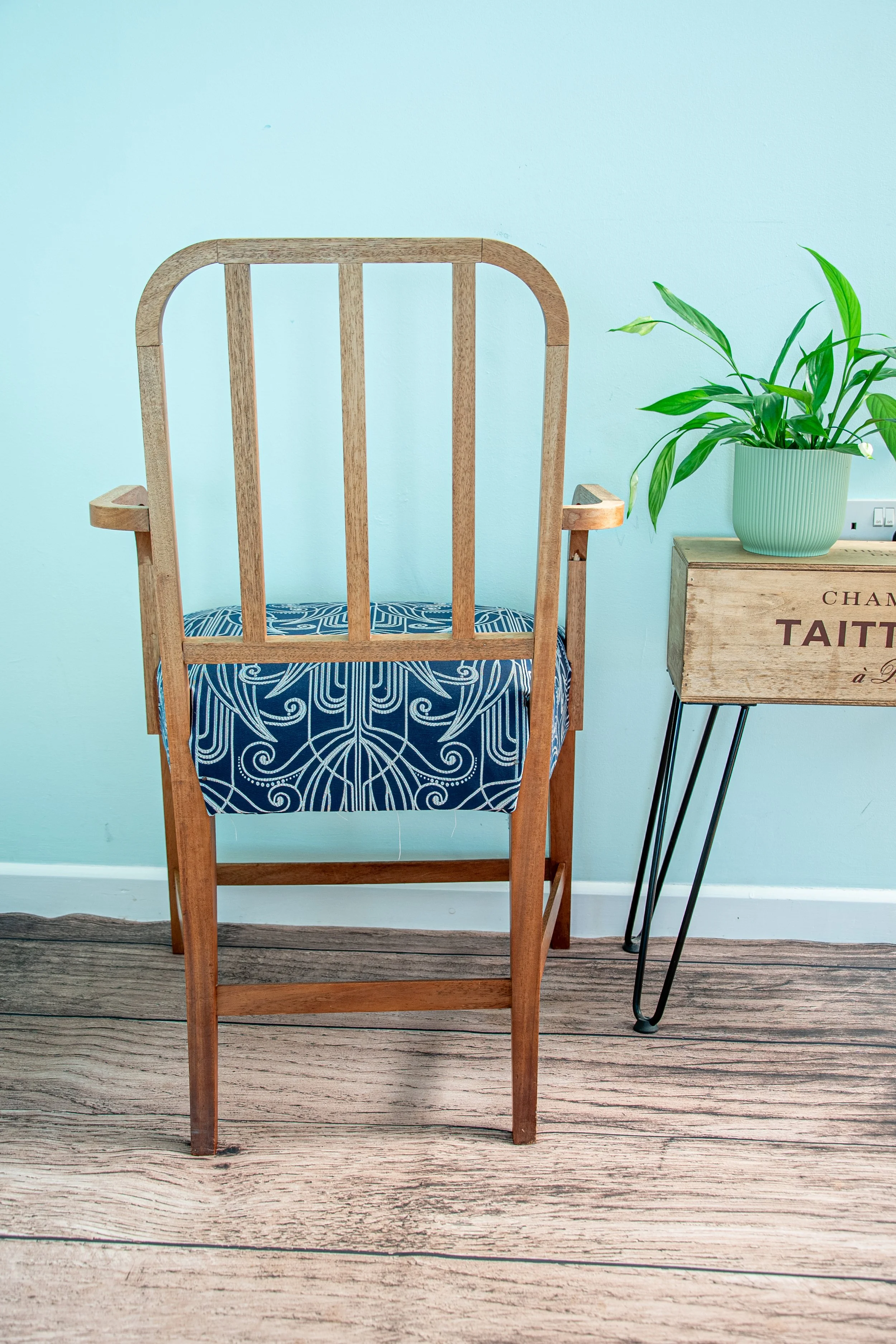 Wooden chair with blue patterned cushion next to a small wooden table with a potted green plant on top, against a light blue wall.