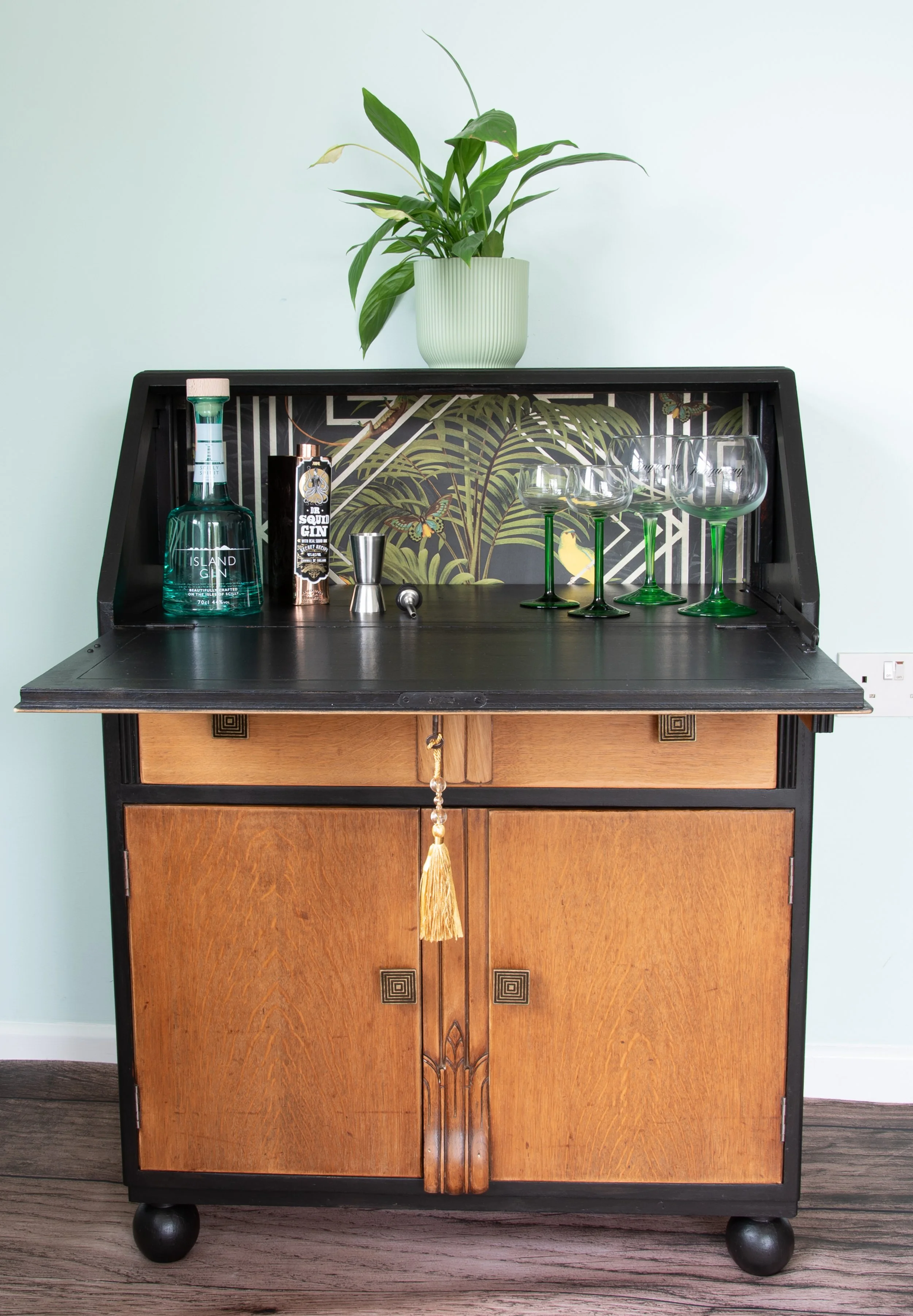 A vintage wooden sideboard with a black top, decorated with a potted plant, liquor bottles, glassware, and cocktail tools, against a white wall.