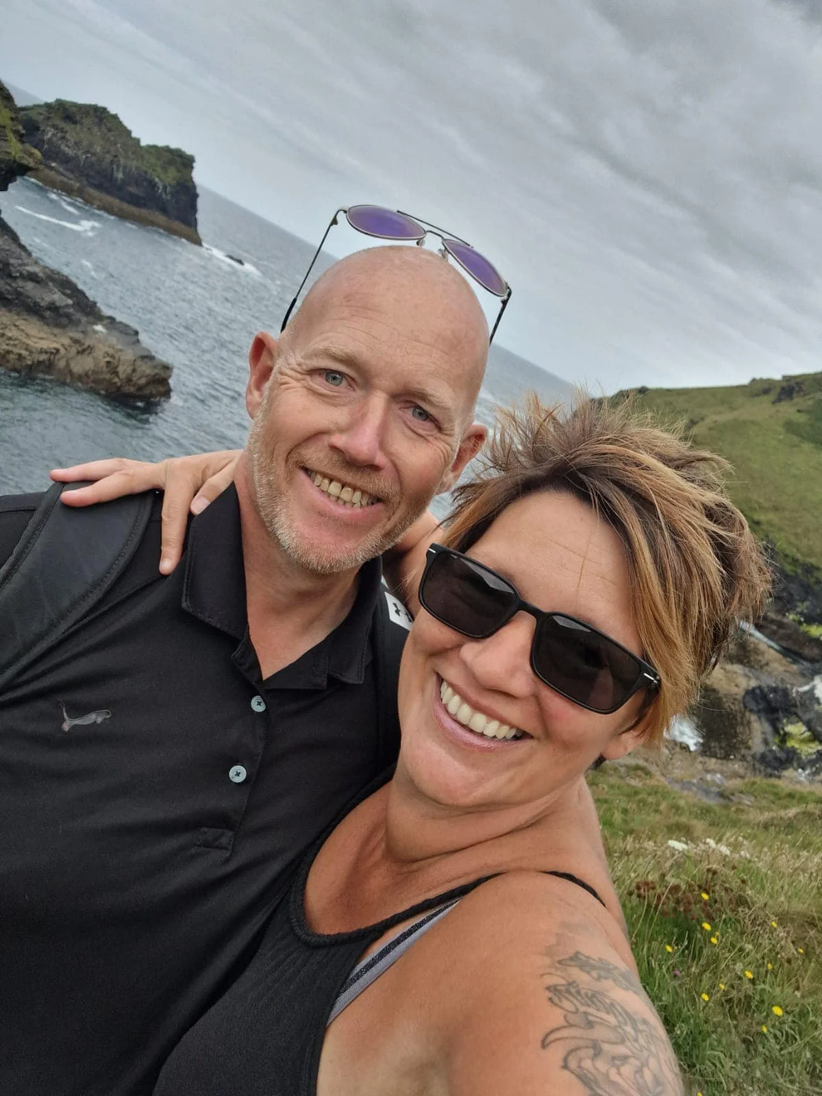 A smiling man and woman taking a selfie near the coast, with rocky cliffs and green hills in the background, overcast sky, the man has a bald head with sunglasses on top, and the woman has short hair with sunglasses.