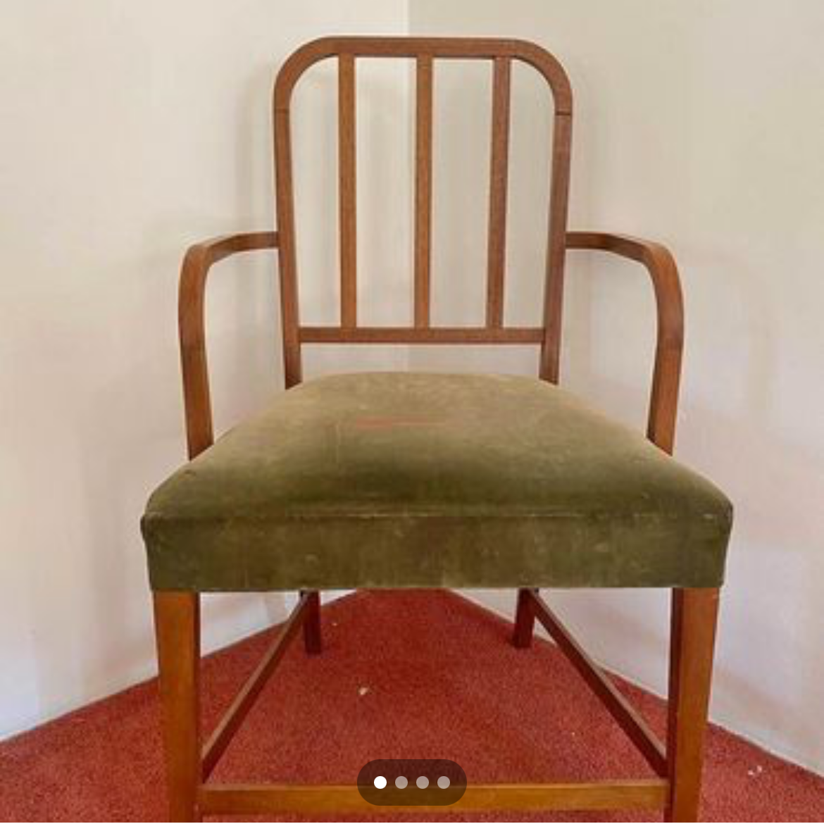 A wooden chair with armrests, a slatted backrest, and a green upholstered seat, placed on a red carpet against a plain light-colored wall.