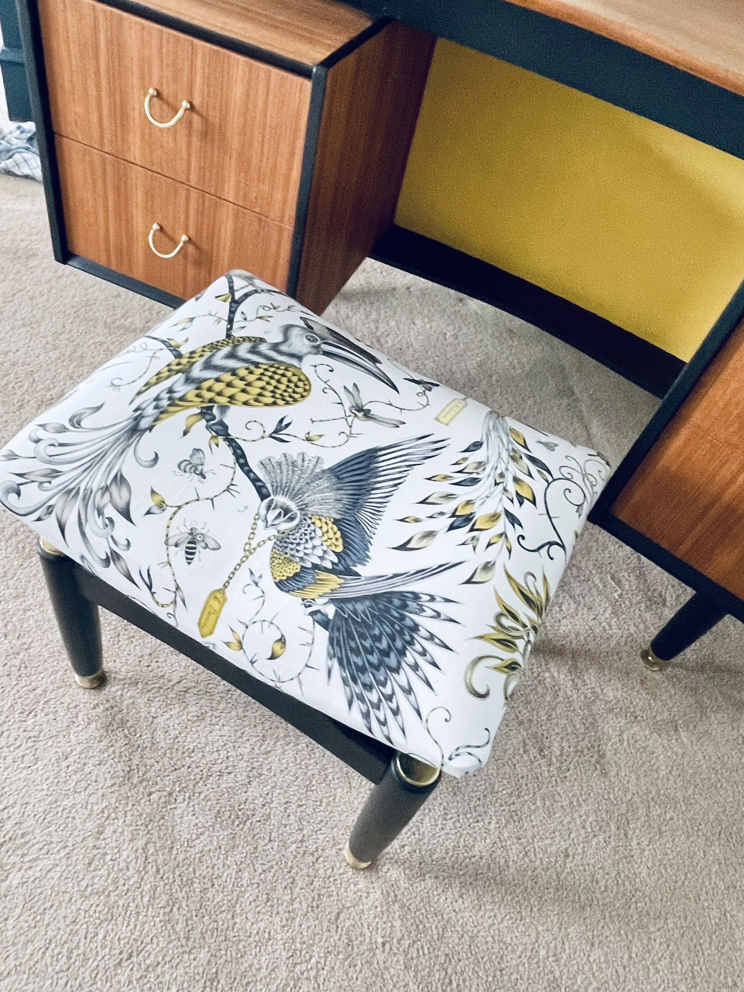 A chair with a black frame, gold accents on the legs, and a cushion featuring a decorative bird design with black, white, and gold colors, placed on a beige carpet.